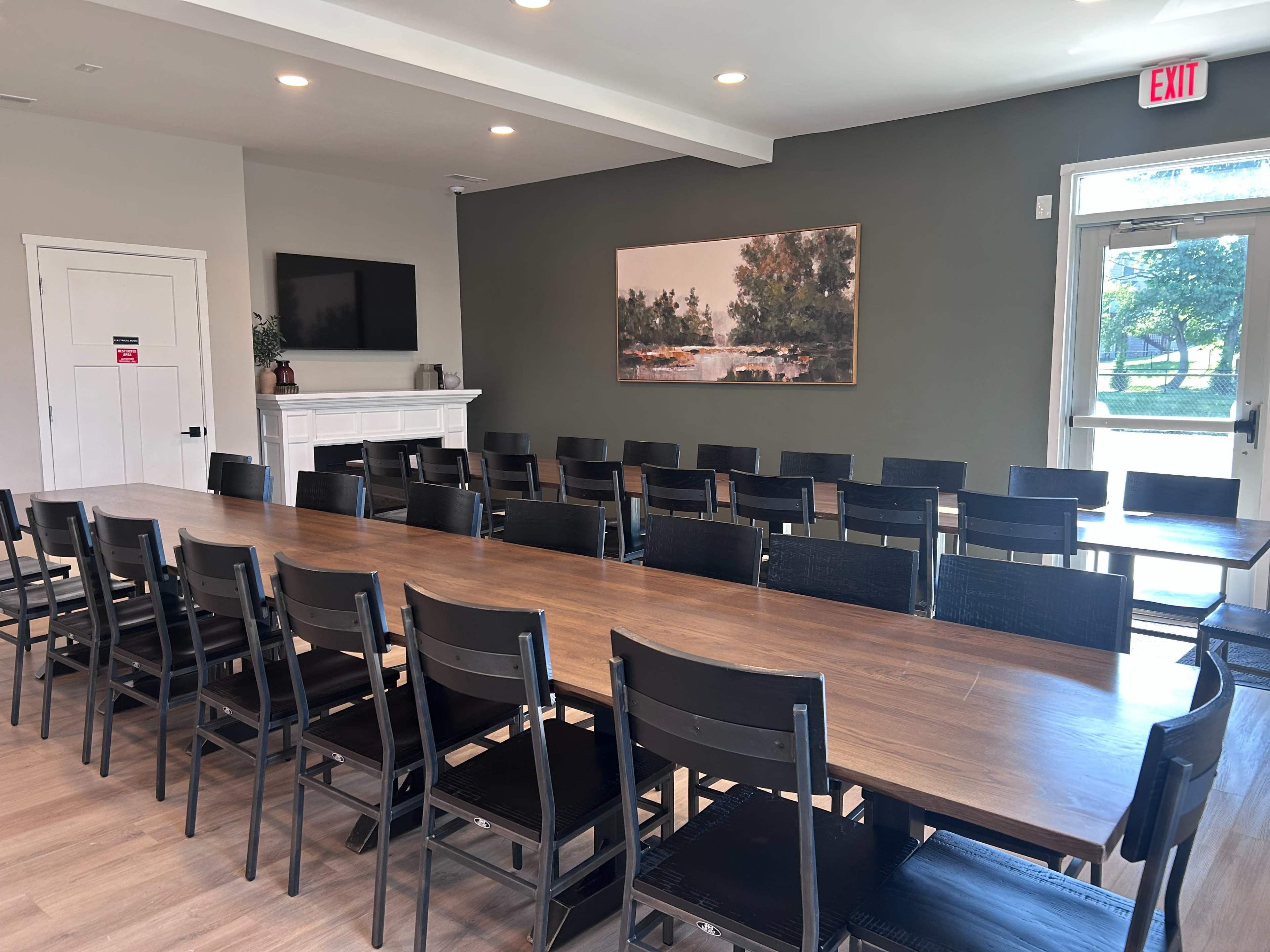 A long wooden table with black chairs is set up in a well-lit meeting room featuring a large painting on the wall and a door leading outside.