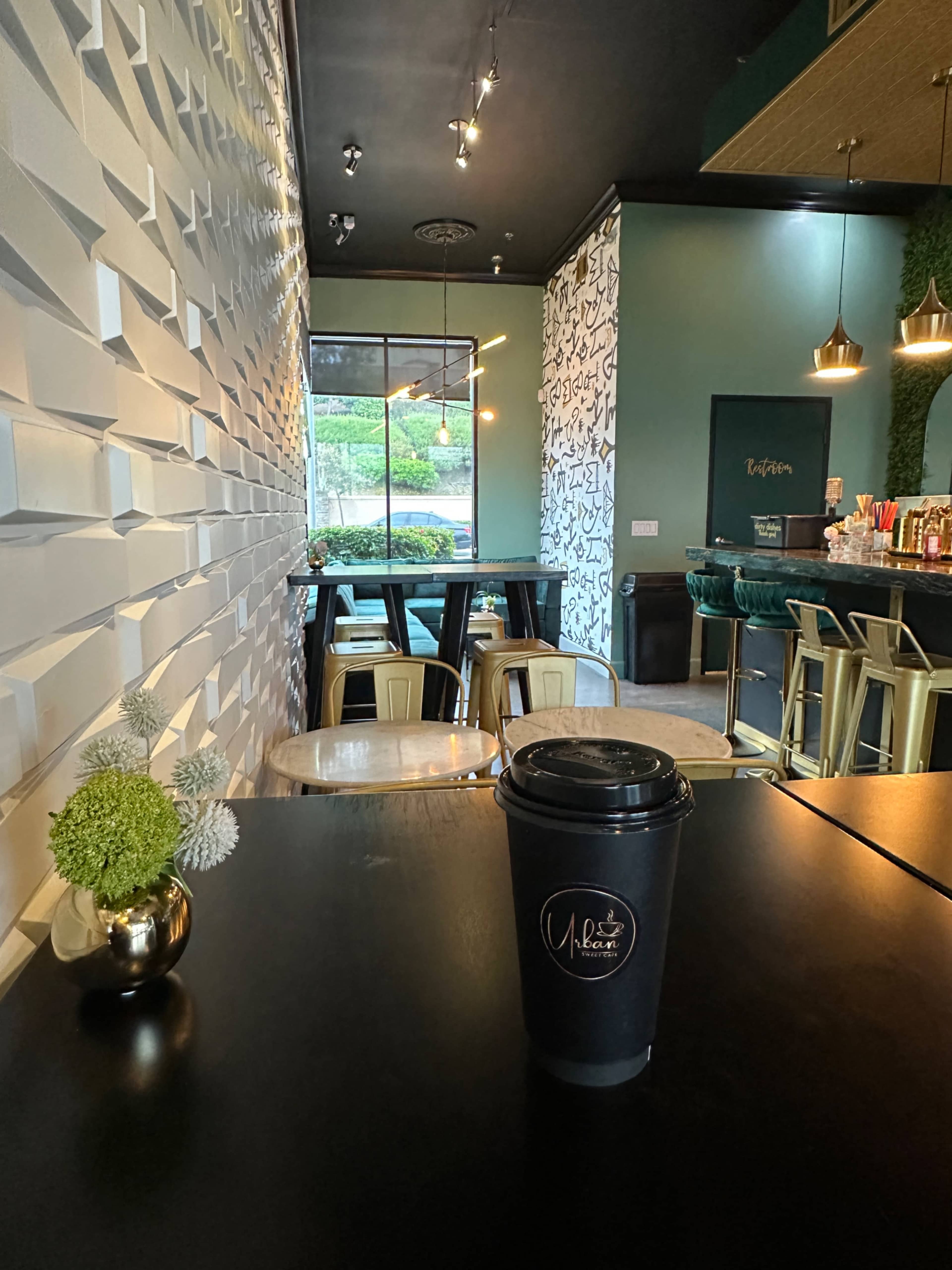 The image features a modern cafe interior with a decorative wall, dark tables, and a takeaway coffee cup prominently displayed in the foreground.