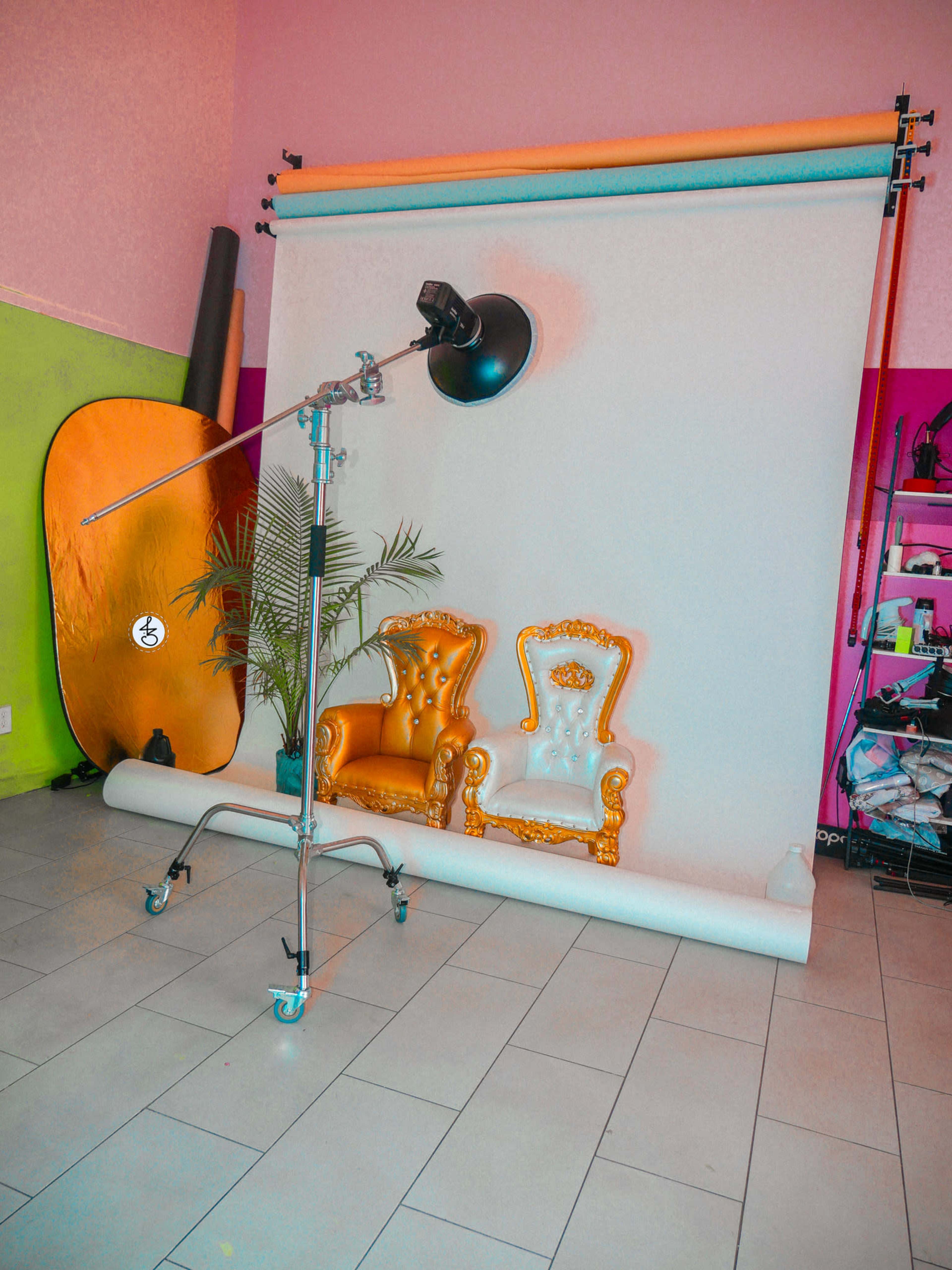 The image shows a photo studio setup with a colorful backdrop, two ornate chairs, a light stand, and a reflector.