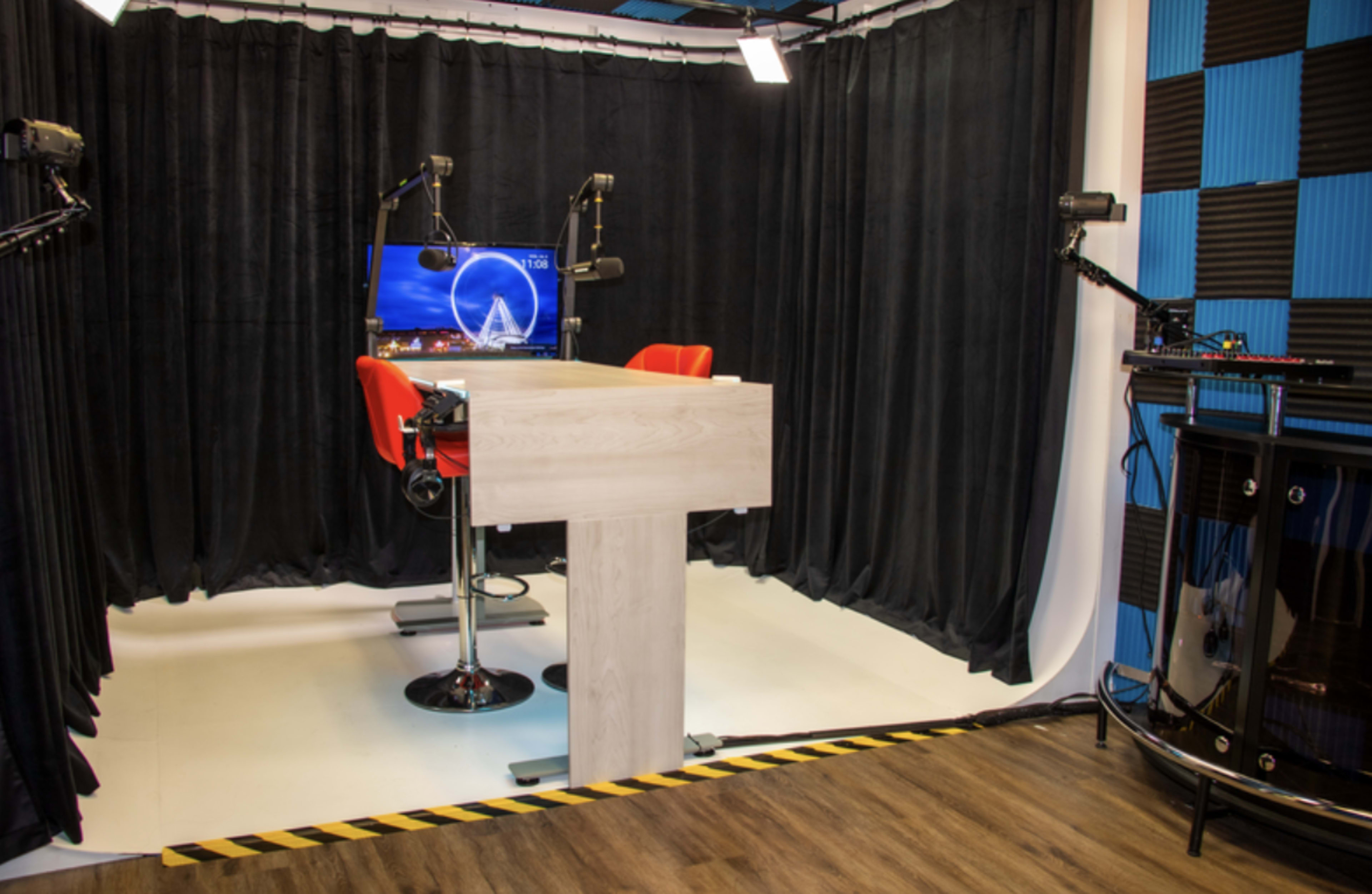 A recording studio with a central table, two red swivel chairs, and a monitor, surrounded by black curtains and a patterned blue wall.