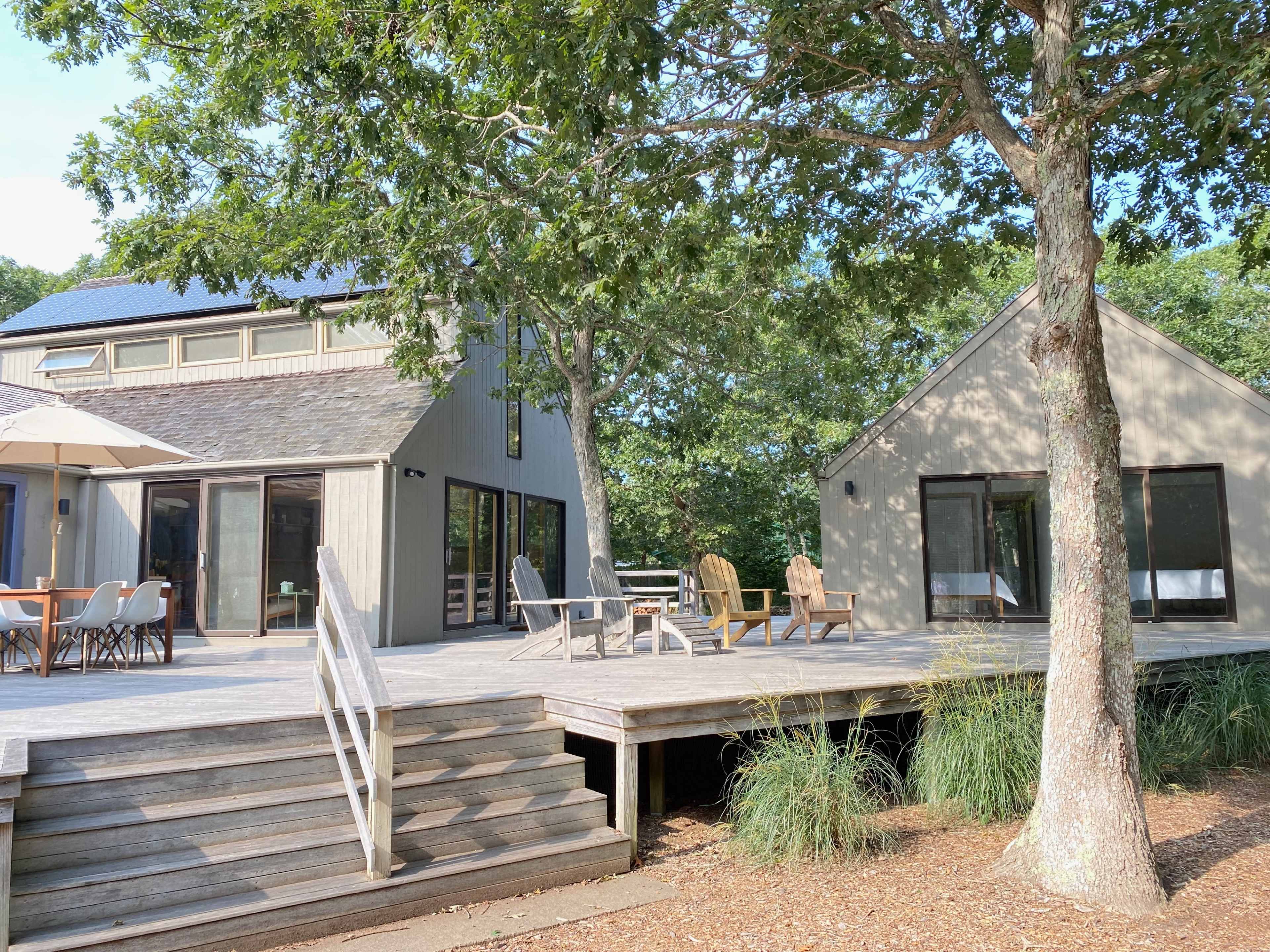 A modern house with a wooden deck, surrounded by trees, features large windows and a shaded patio area.