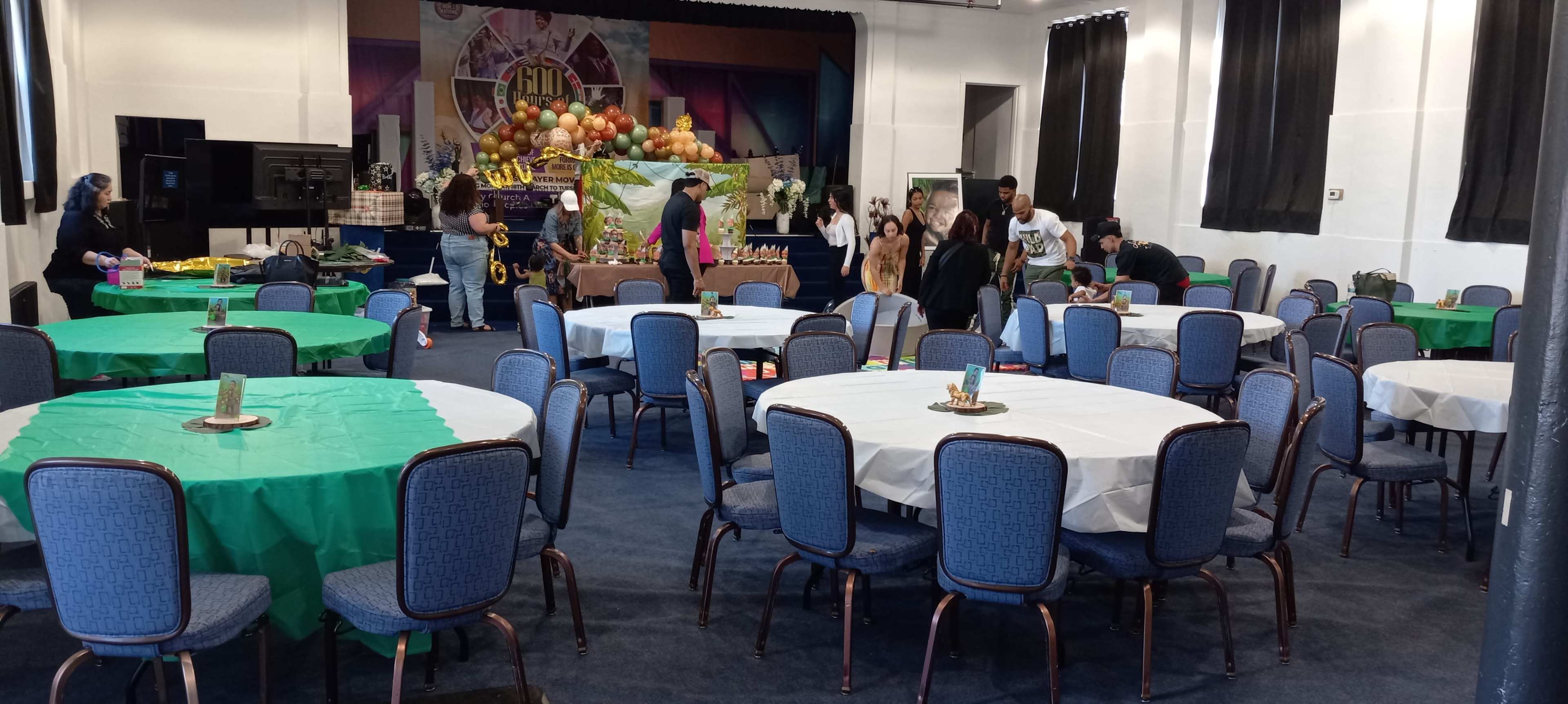 The image shows a large event space with several round tables covered in white tablecloths and green overlays, where people are arranging decorations and preparing for an event.