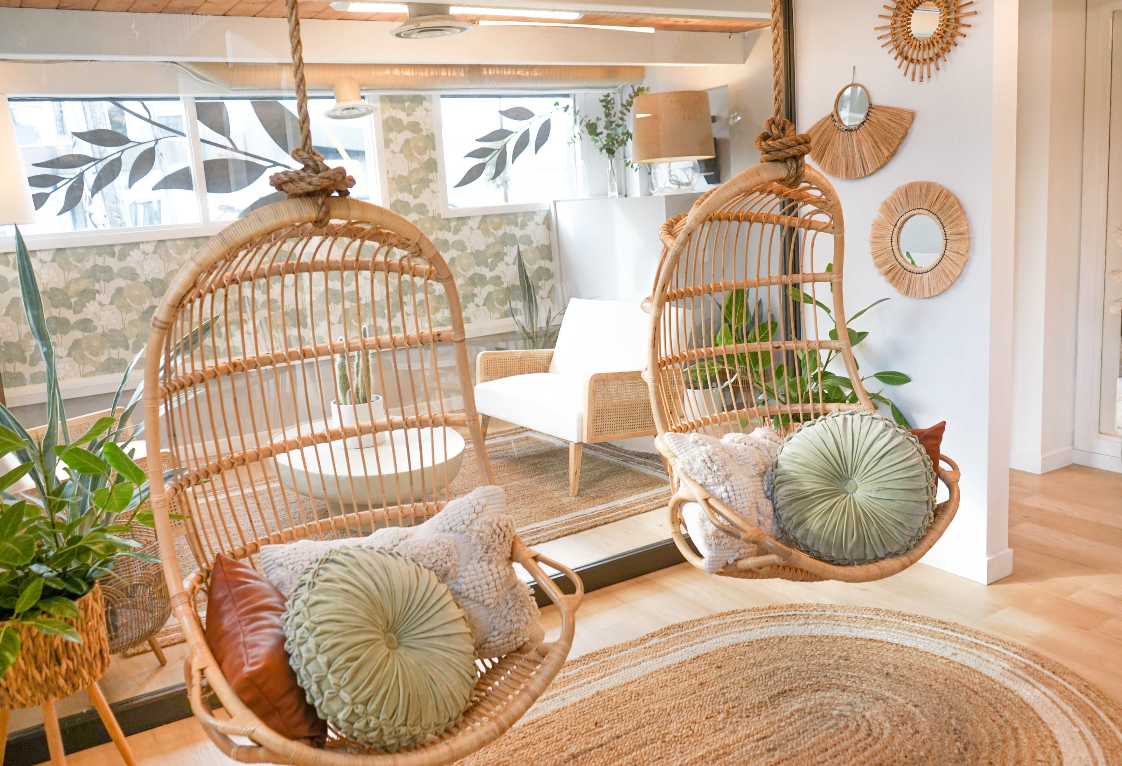 Two woven hanging chairs with cushions, positioned in a bright room decorated with plants and mirrors.