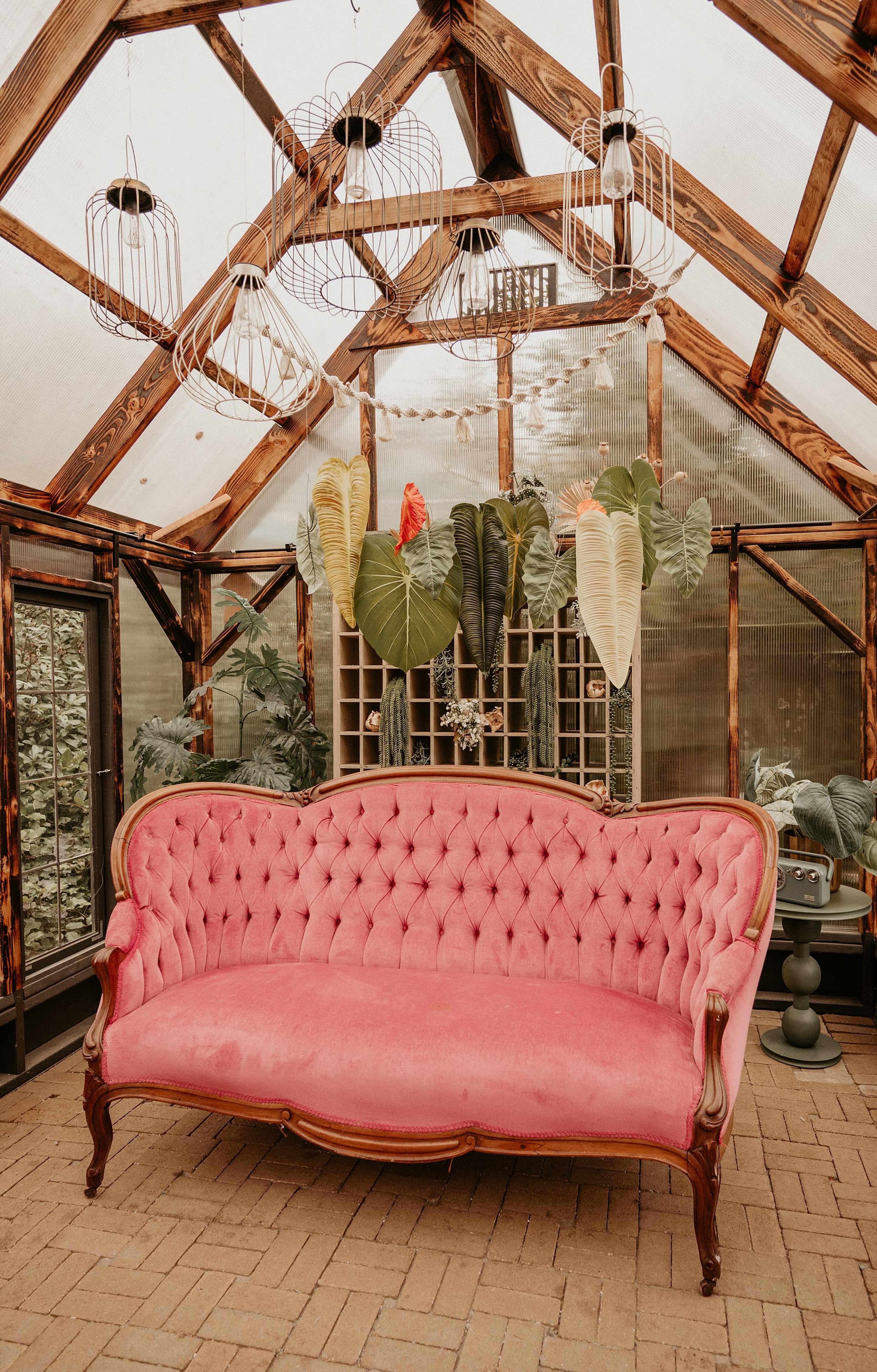 A pink tufted sofa is positioned inside a greenhouse with large plants in the background and decorative hanging lights above.