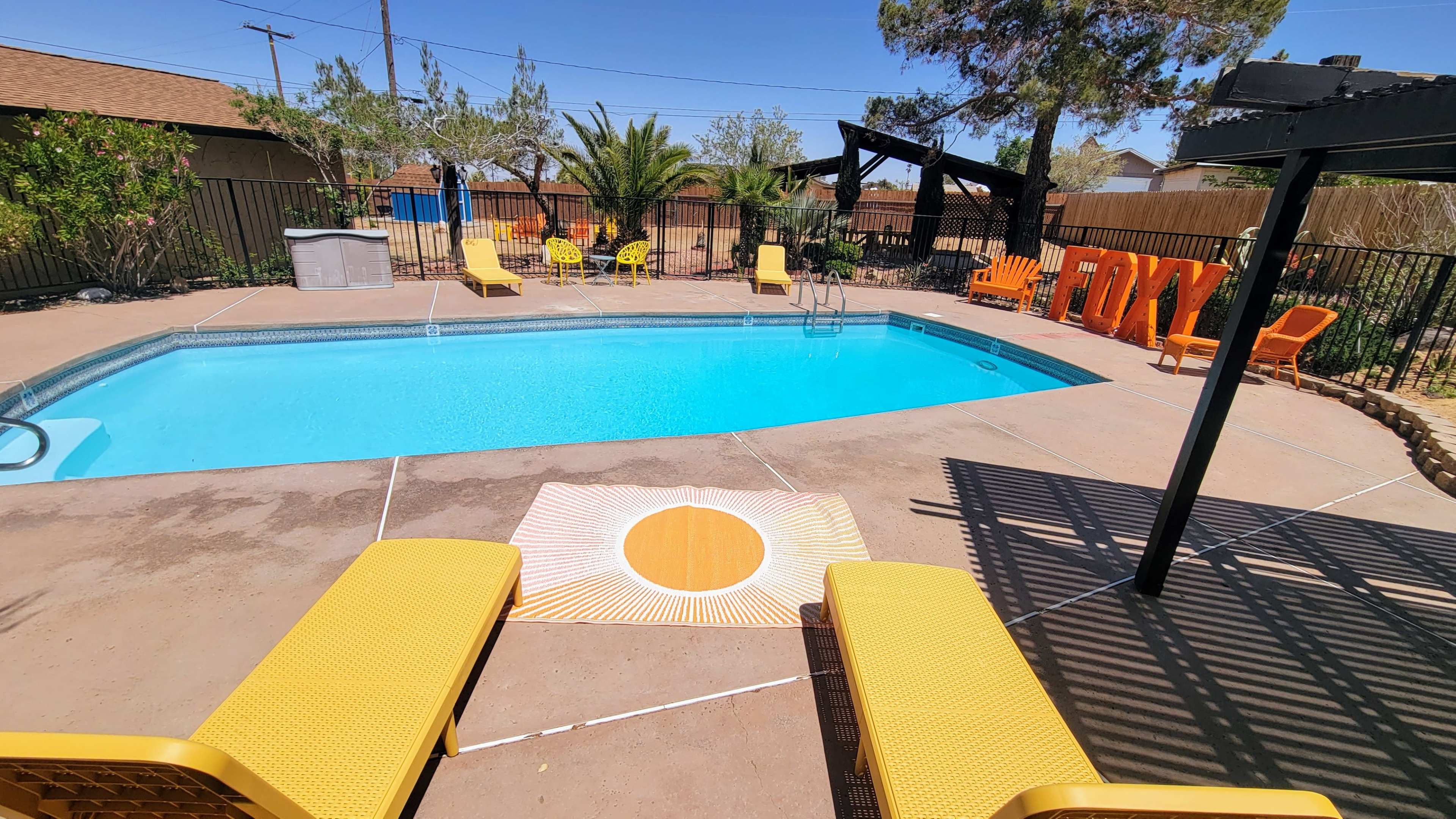 A bright blue swimming pool surrounded by yellow lounge chairs and a sun-patterned mat in a landscaped backyard.