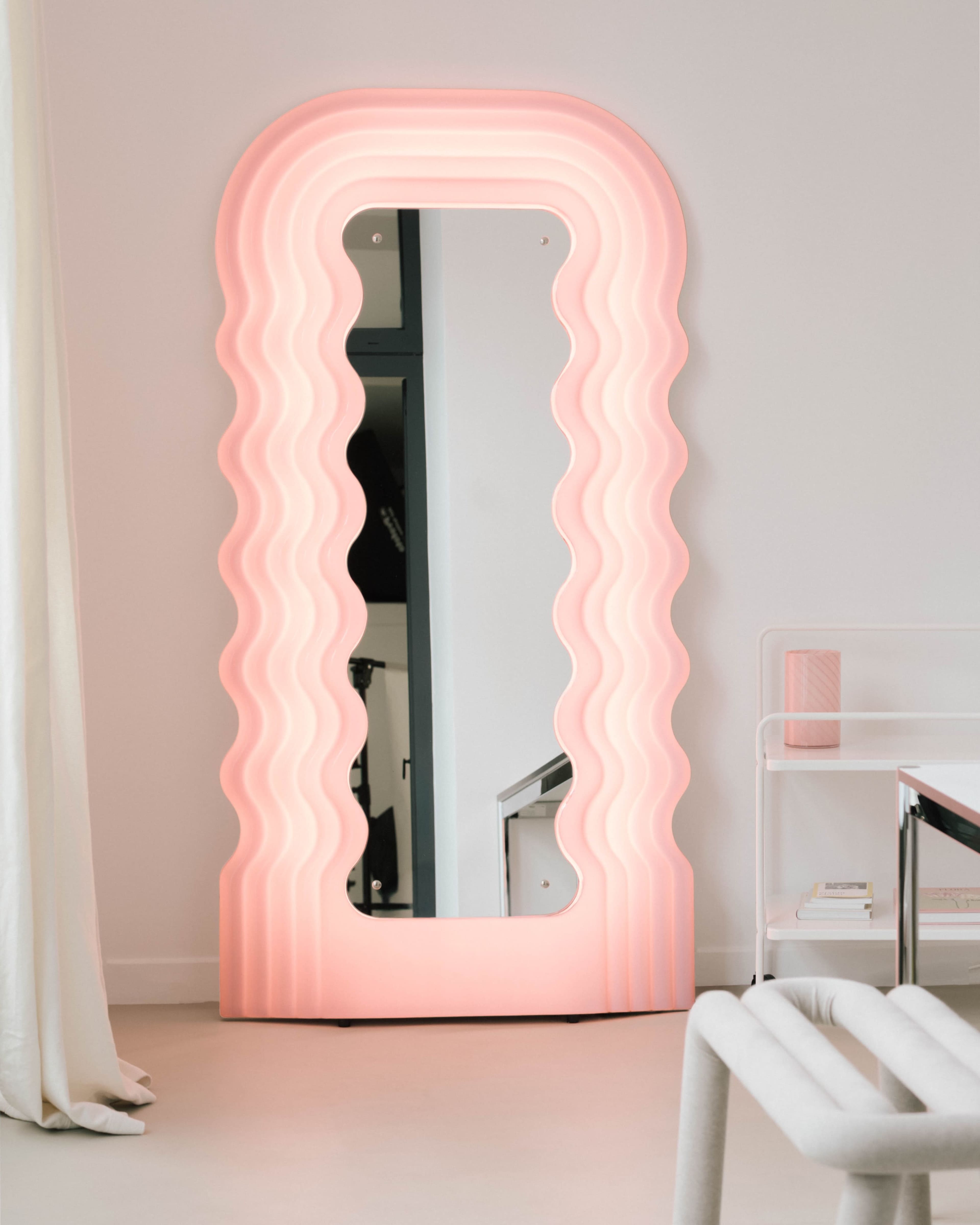 A decorative mirror with a wavy pink frame stands against a white wall in a modern interior space.