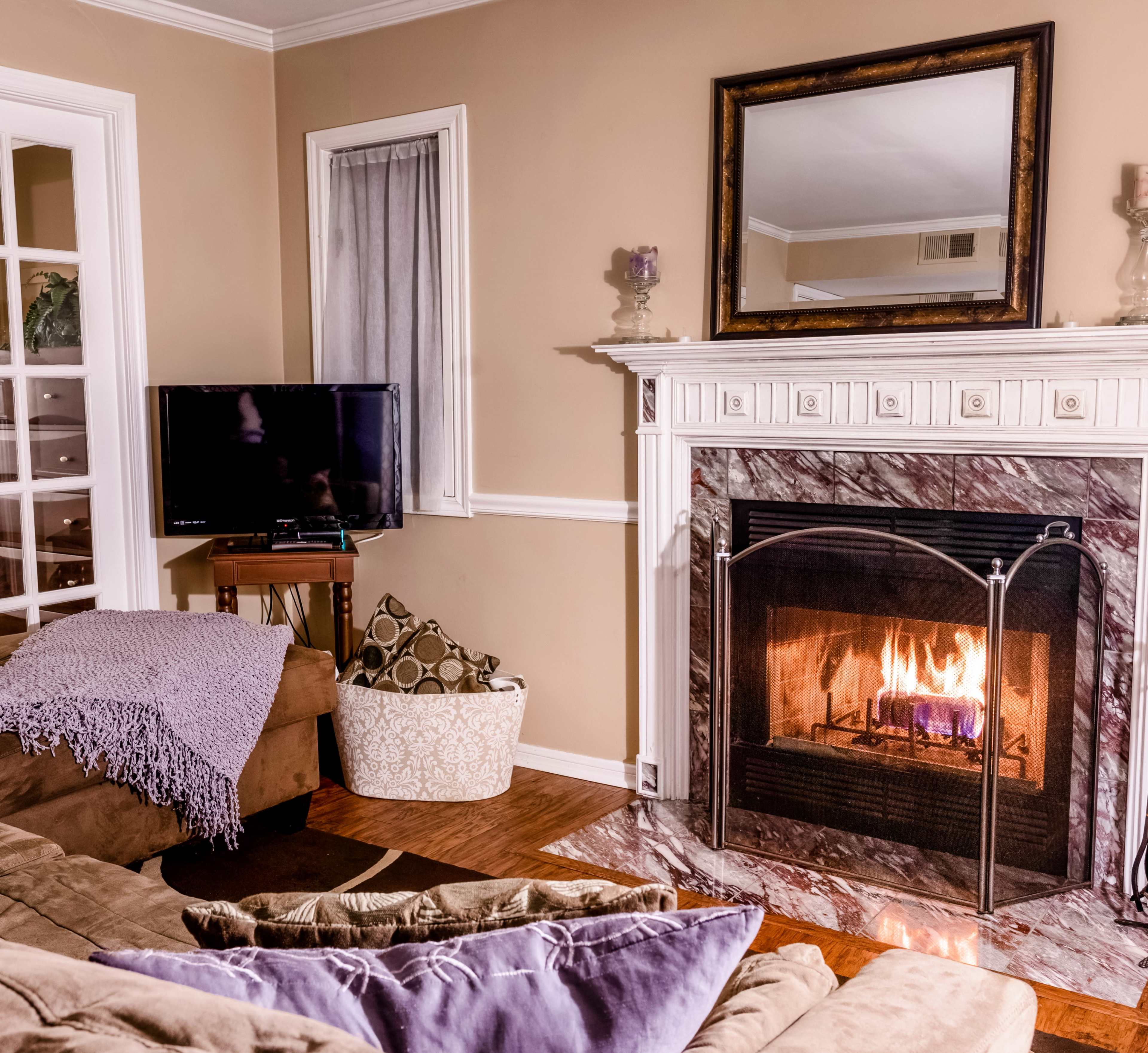 A cozy living room features a fireplace with a flickering flame, a television on a stand, and a brown sectional couch adorned with purple pillows.