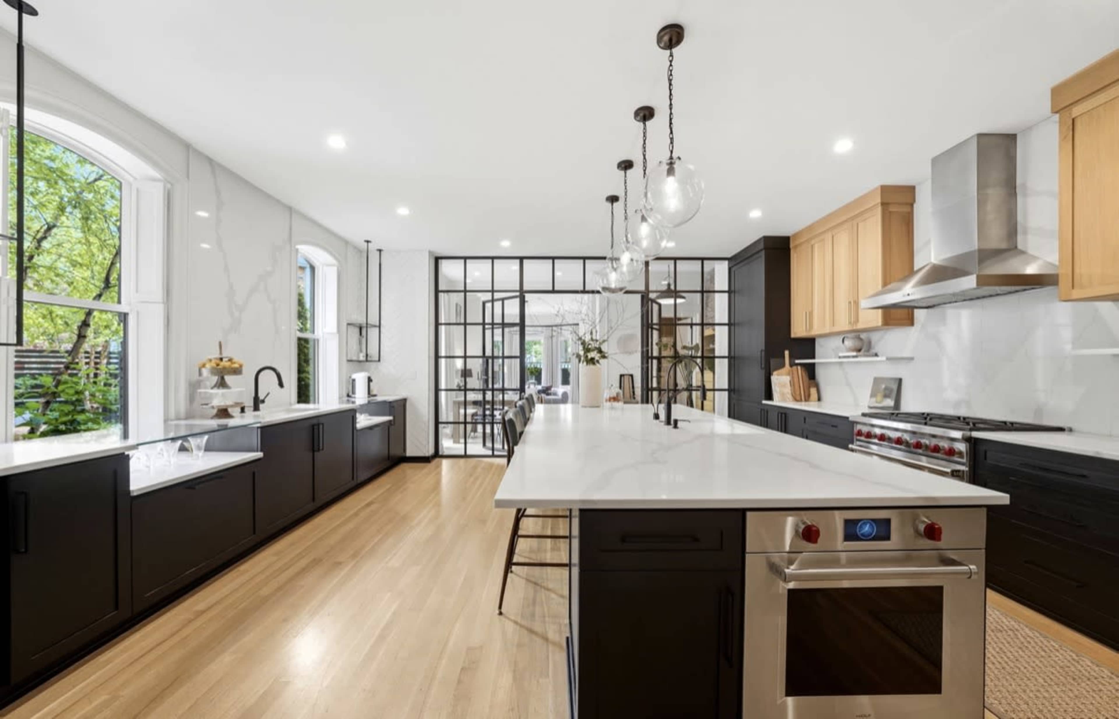 The kitchen features a large central island with a white countertop, black cabinetry, and stainless steel appliances, surrounded by windows and a glass-paneled wall dividing the space.