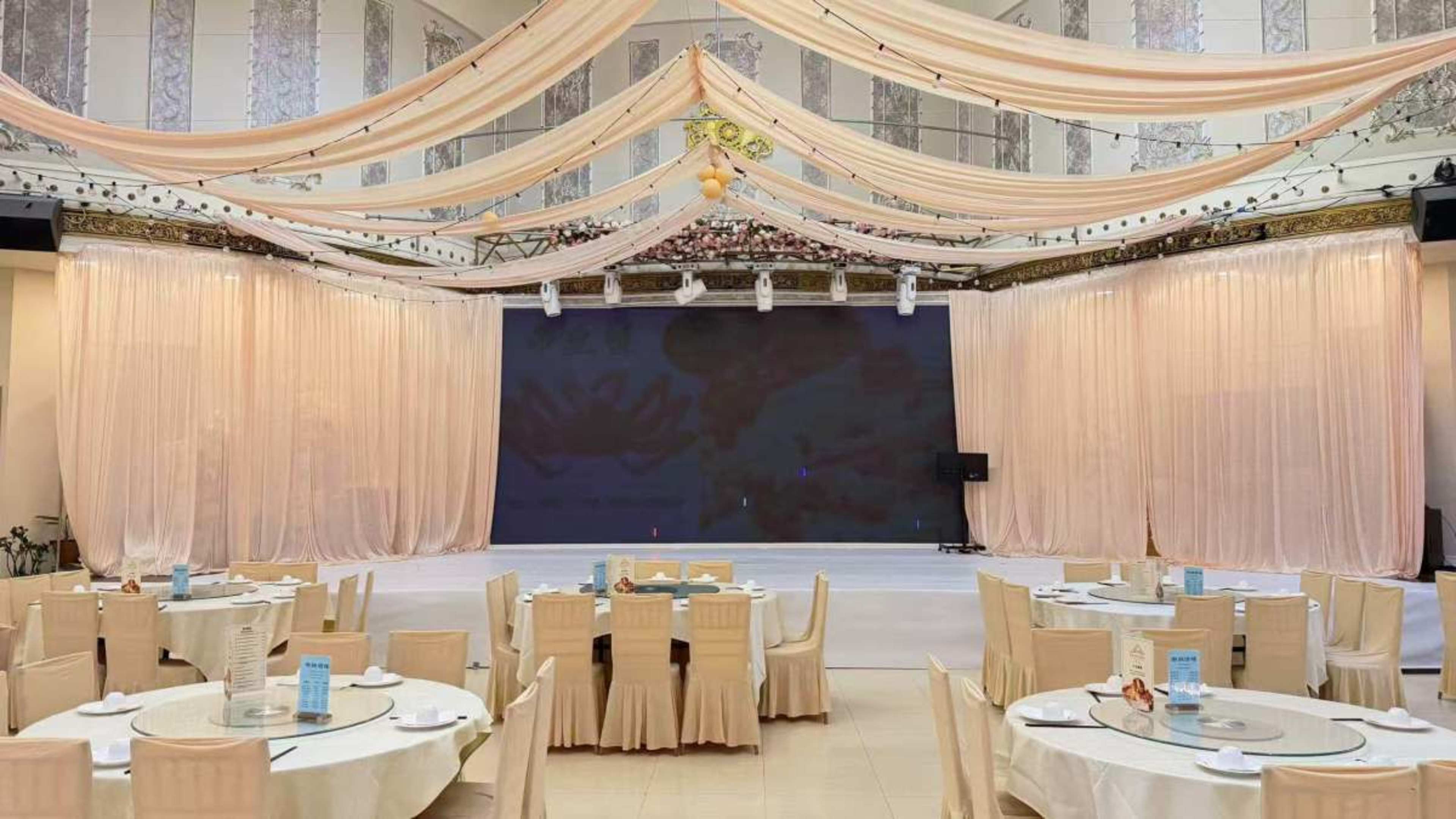 The image shows a spacious event hall with elegantly draped fabric, round tables set for dining, and a stage area in the background.