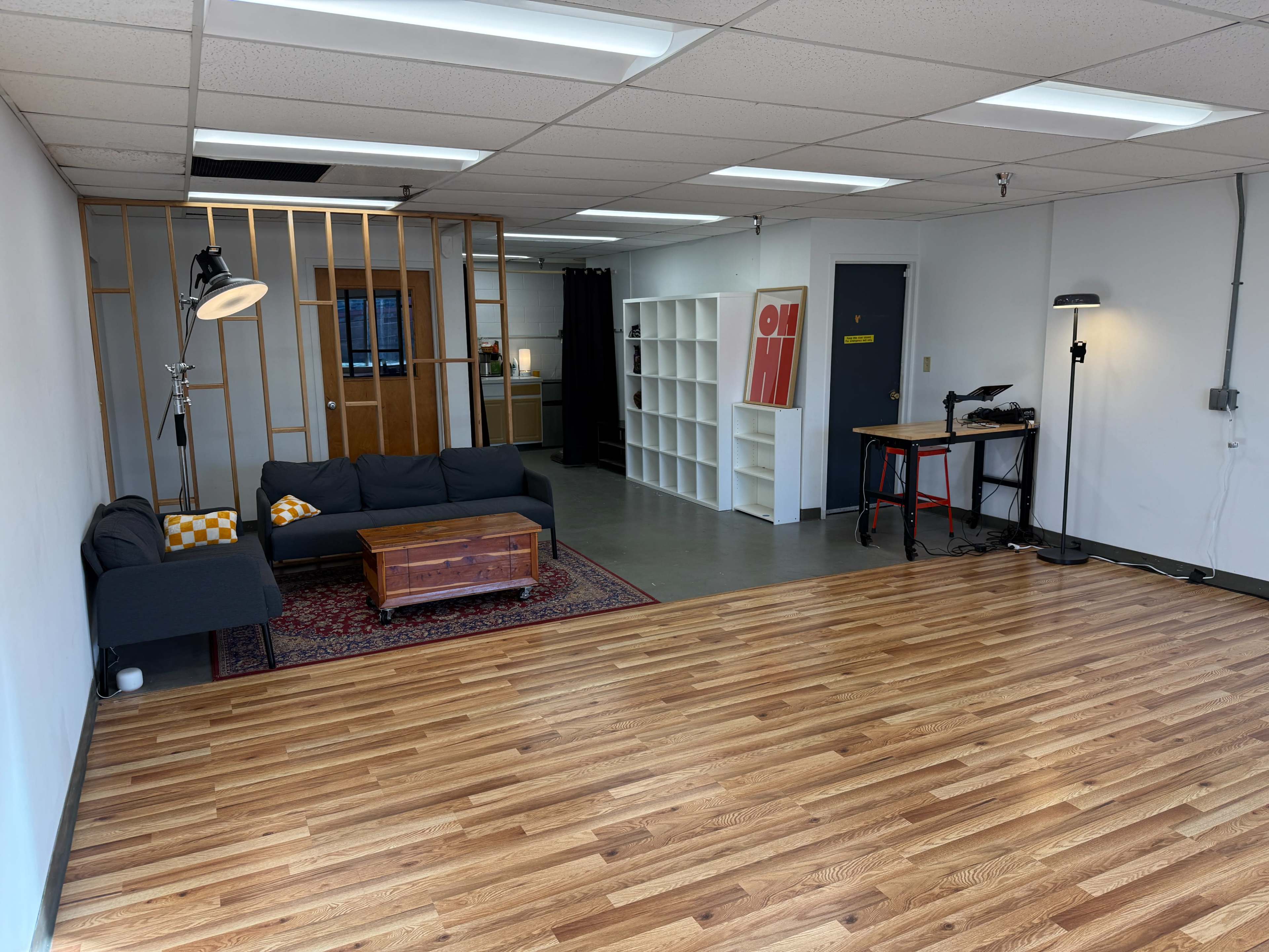 The image shows a spacious room with wooden flooring, featuring a dark couch, a coffee table, and a bookshelf against one wall, with a partition and a lamp near the center.
