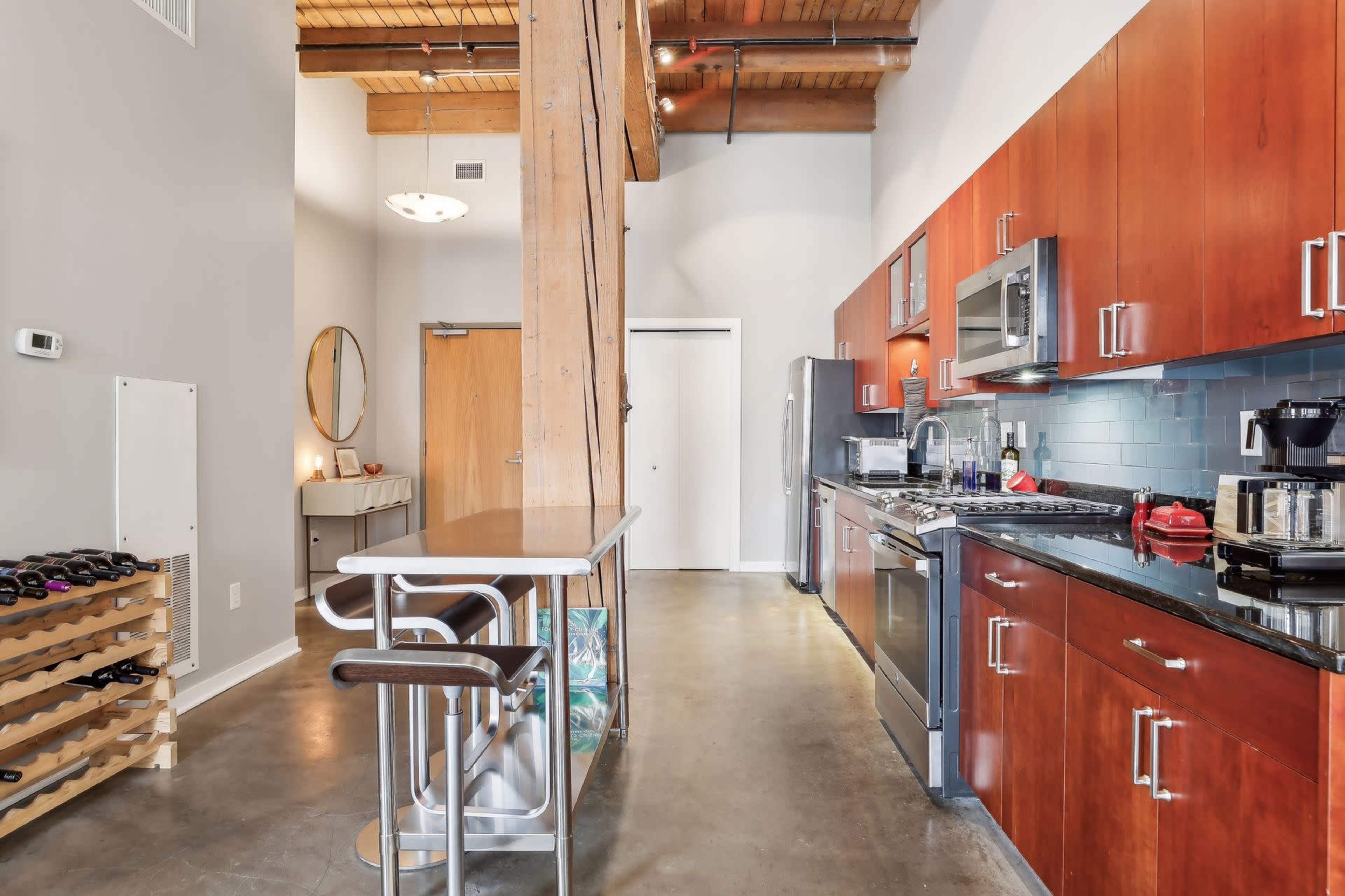 The image shows a modern kitchen and dining area with wooden beams, stainless steel appliances, and a bar table with two stools.