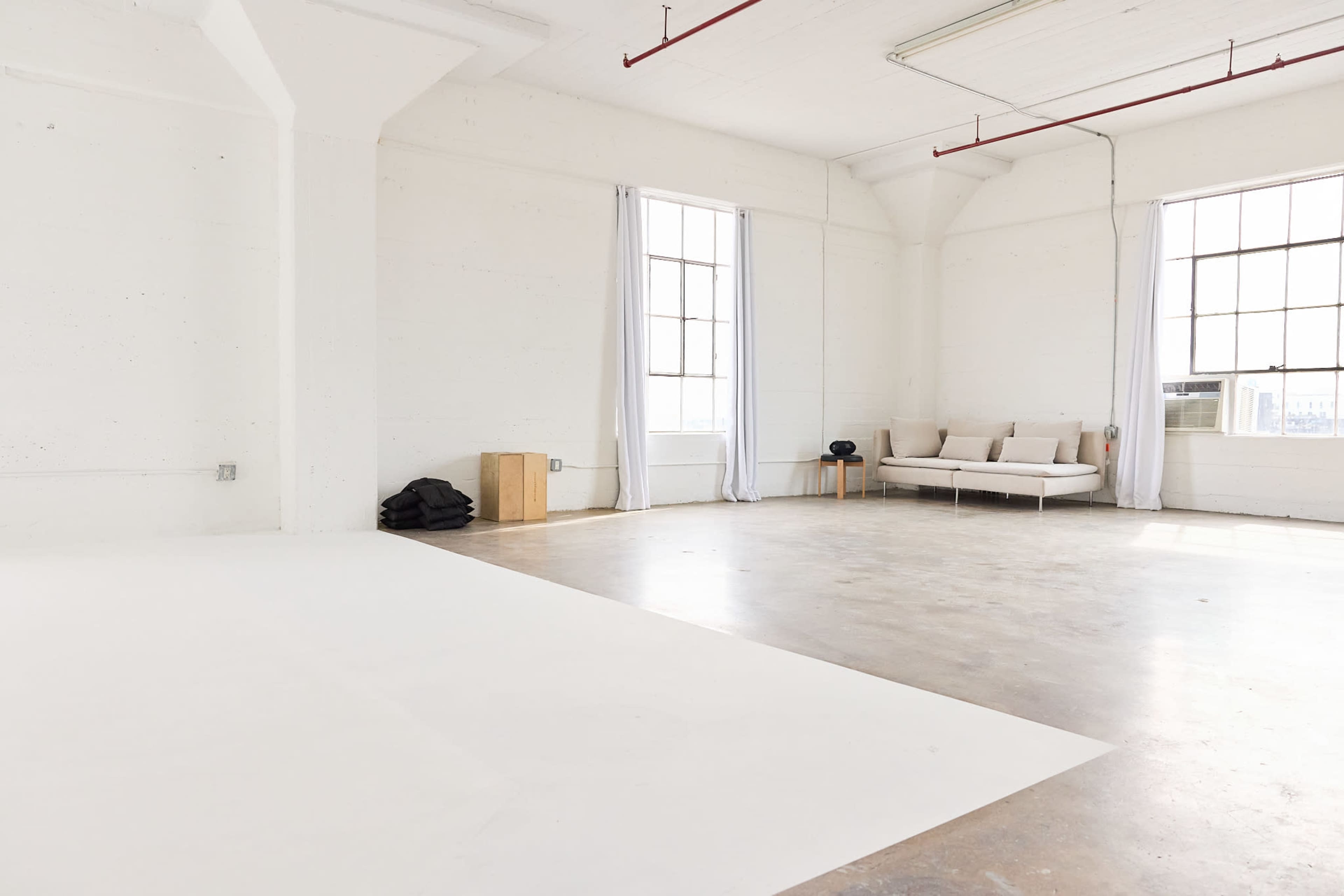 The image displays a spacious, minimally decorated interior with white walls, large windows, and a light-colored sofa.