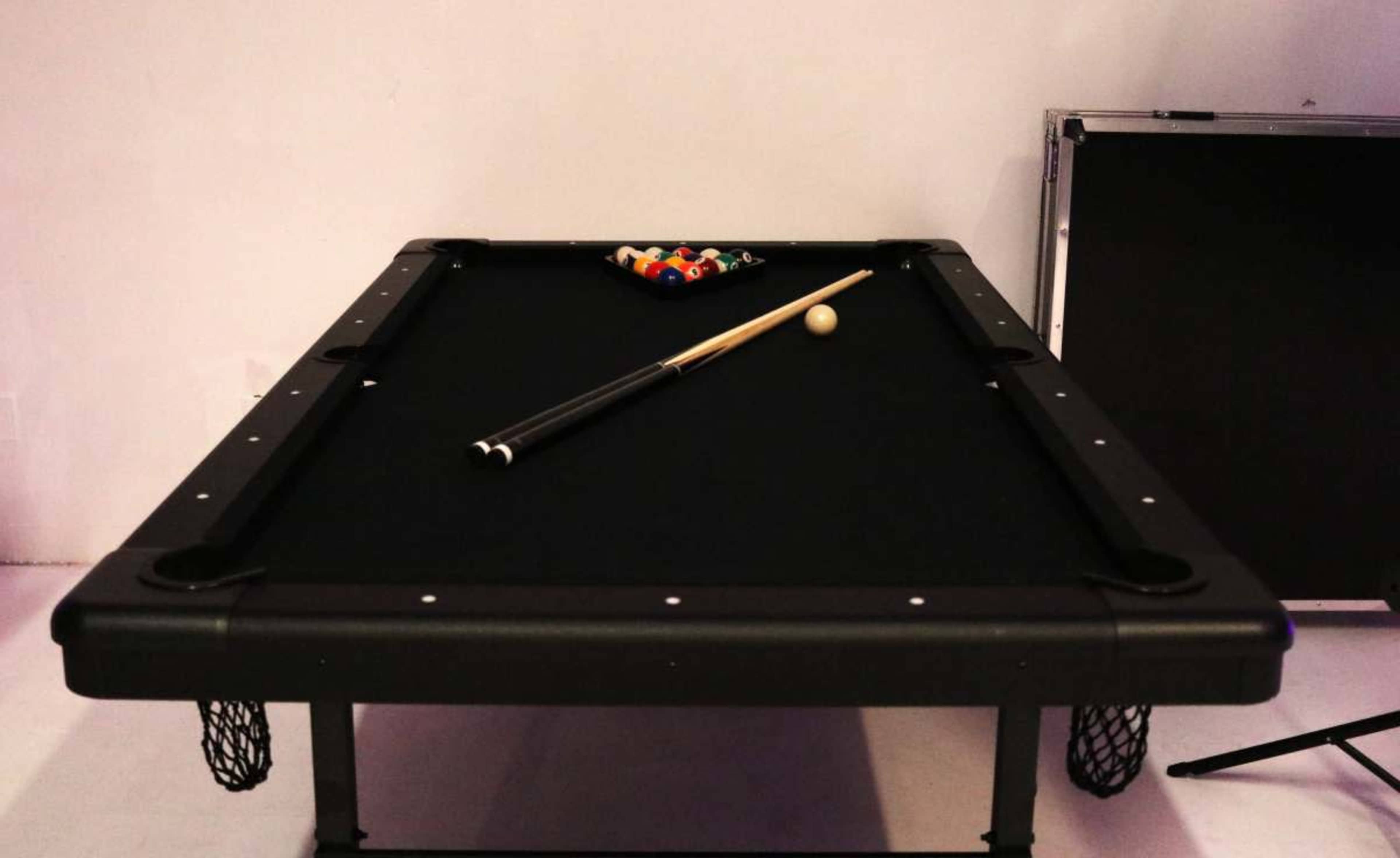 A black billiards table is set up with a cue stick resting on it and a rack of colored balls arranged at one end.