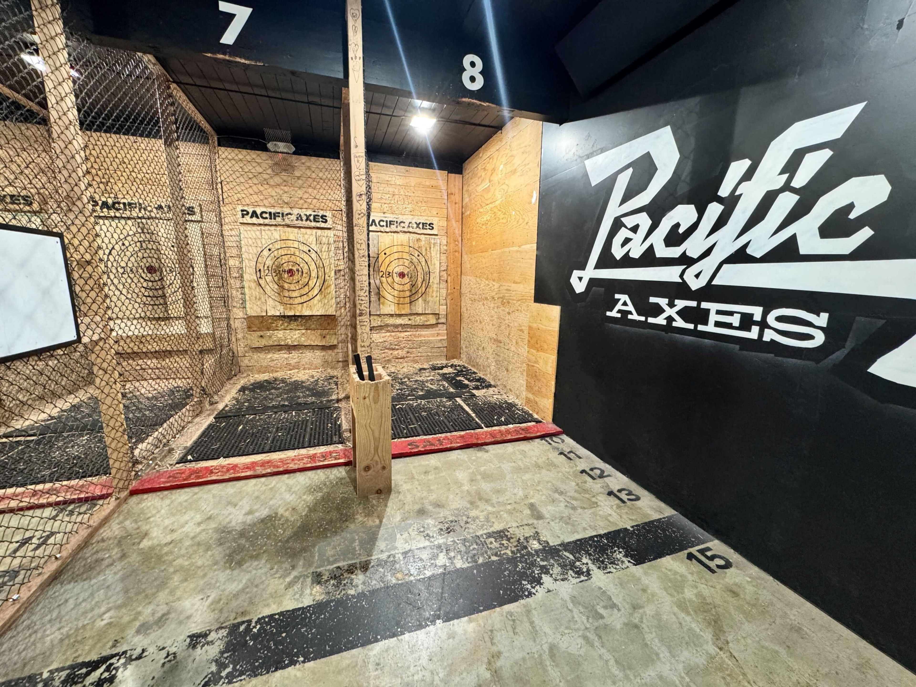 The image shows an indoor axe throwing facility with two target lanes, surrounded by protective fencing and marked floor lines.