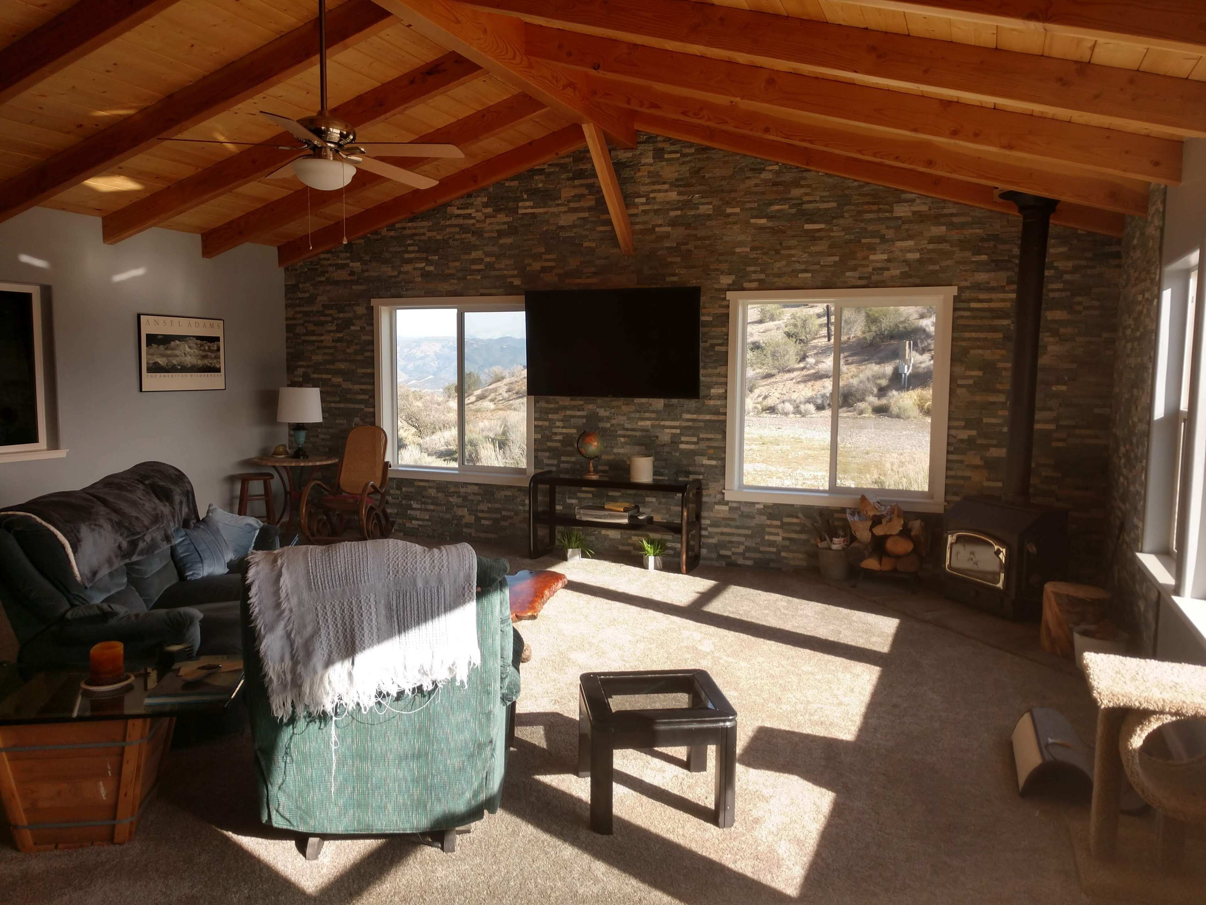 The living room features a stone wall, large windows with a view, and a wood-burning stove, all under a sloped wooden ceiling.