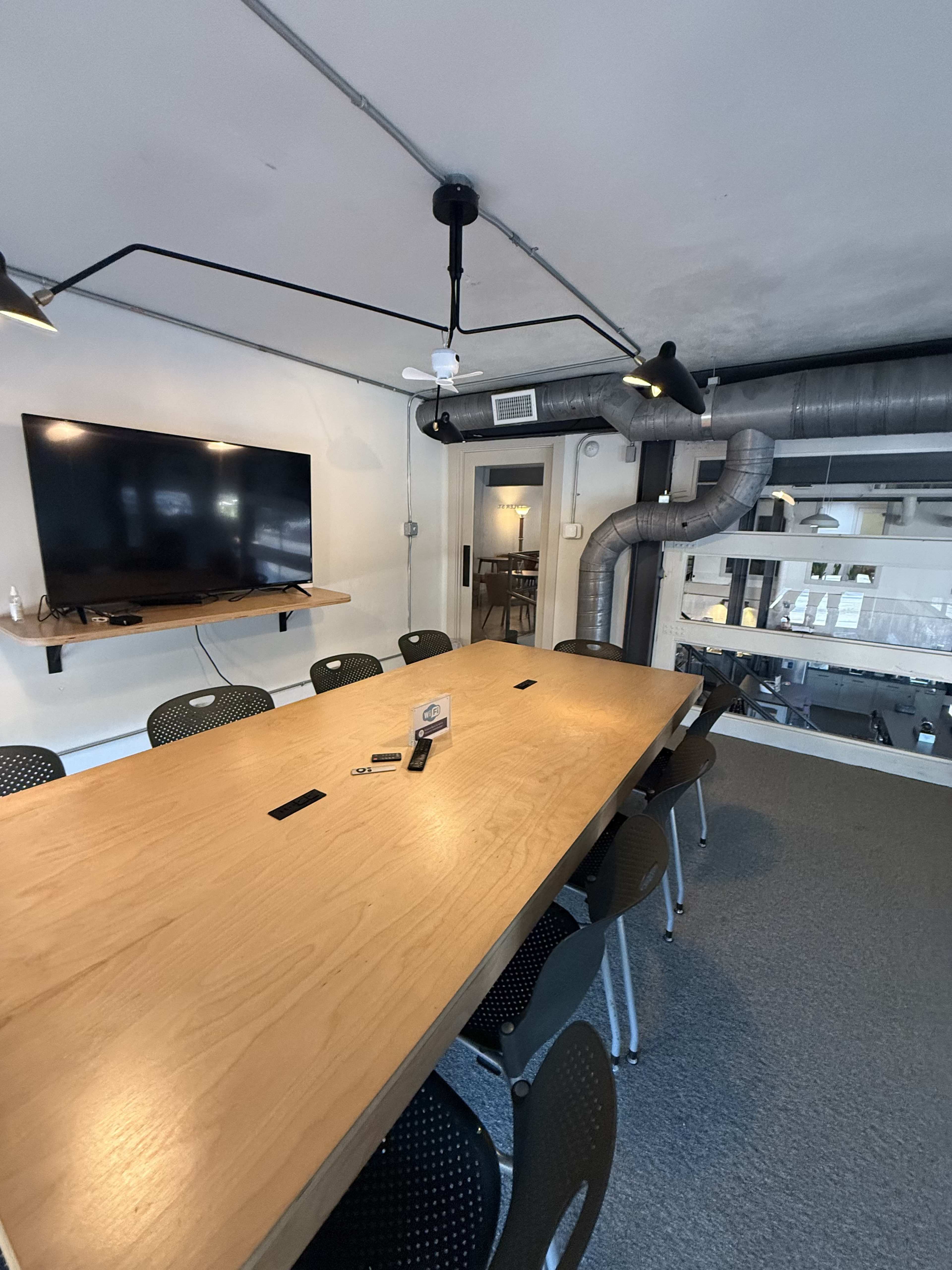 The image shows a modern conference room featuring a large wooden table surrounded by black chairs, with a wall-mounted television and exposed ducts on the ceiling.