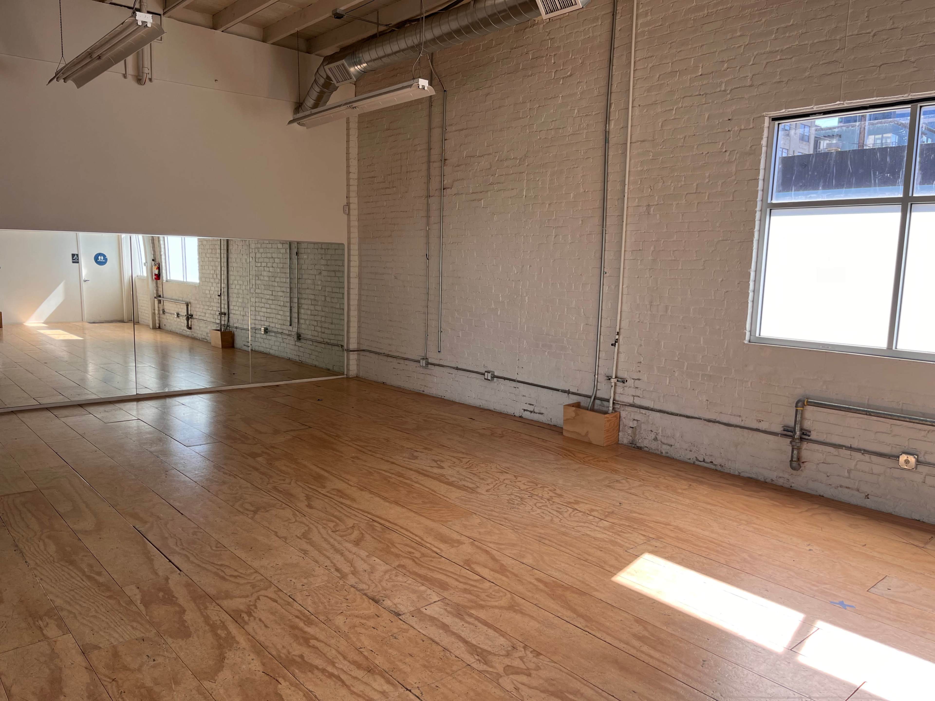 A spacious room with wooden flooring and a large mirror on one wall, illuminated by natural light from a window.