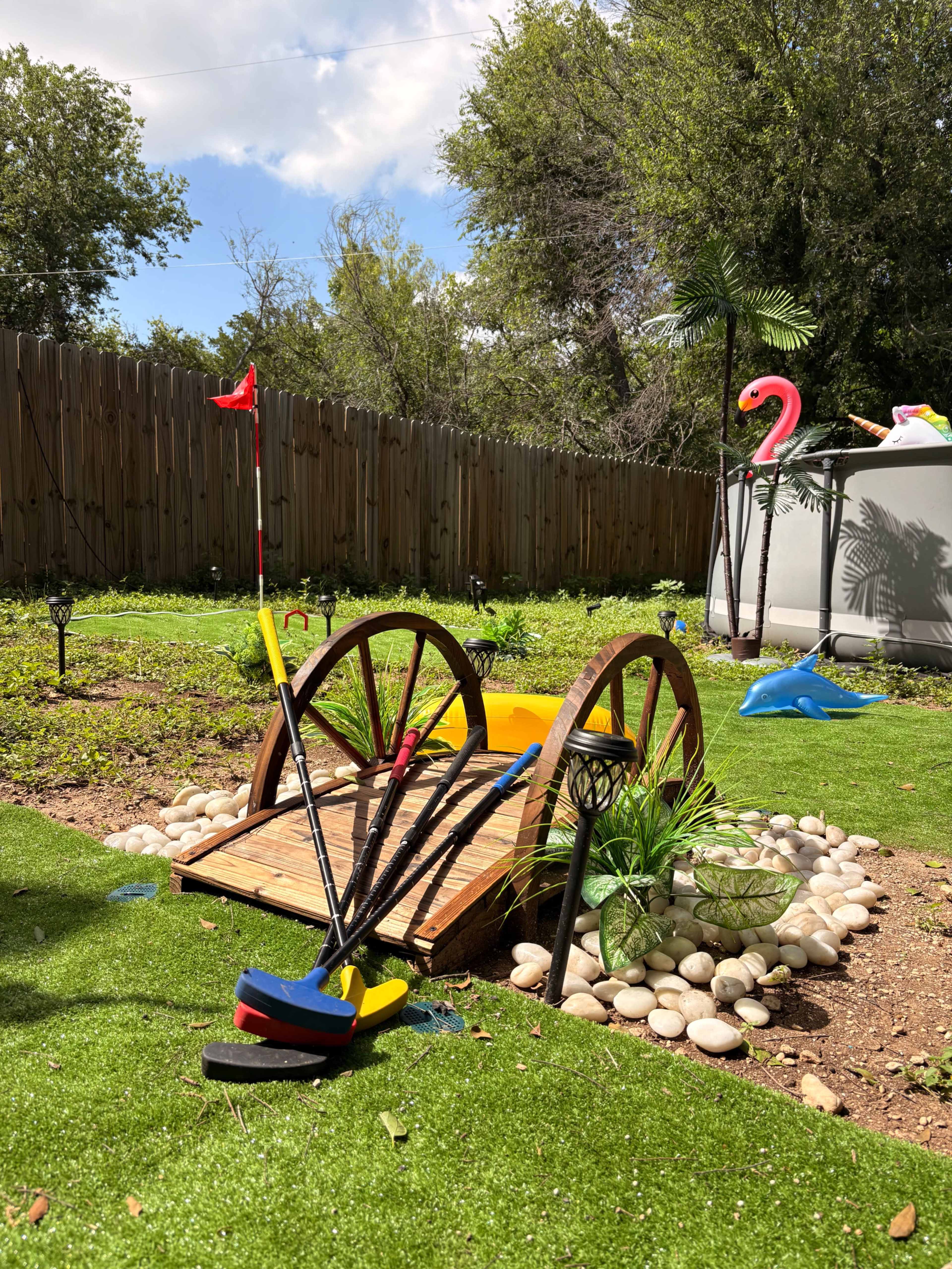 A wooden bridge crosses over a small area of artificial grass adorned with colorful garden decorations and plants.