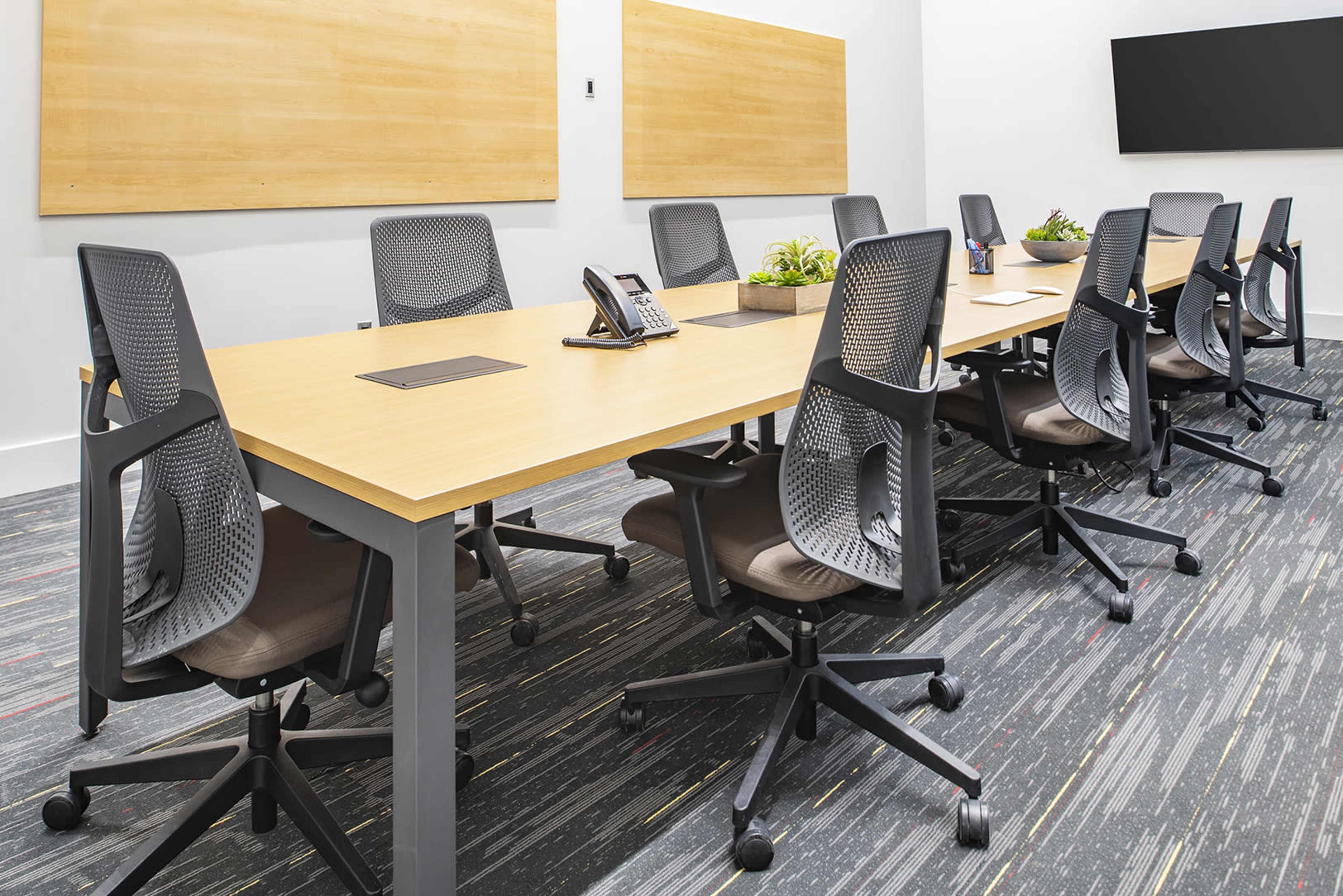 A modern conference room features a long table surrounded by rolling office chairs, with wooden wall panels and a large screen on one side.