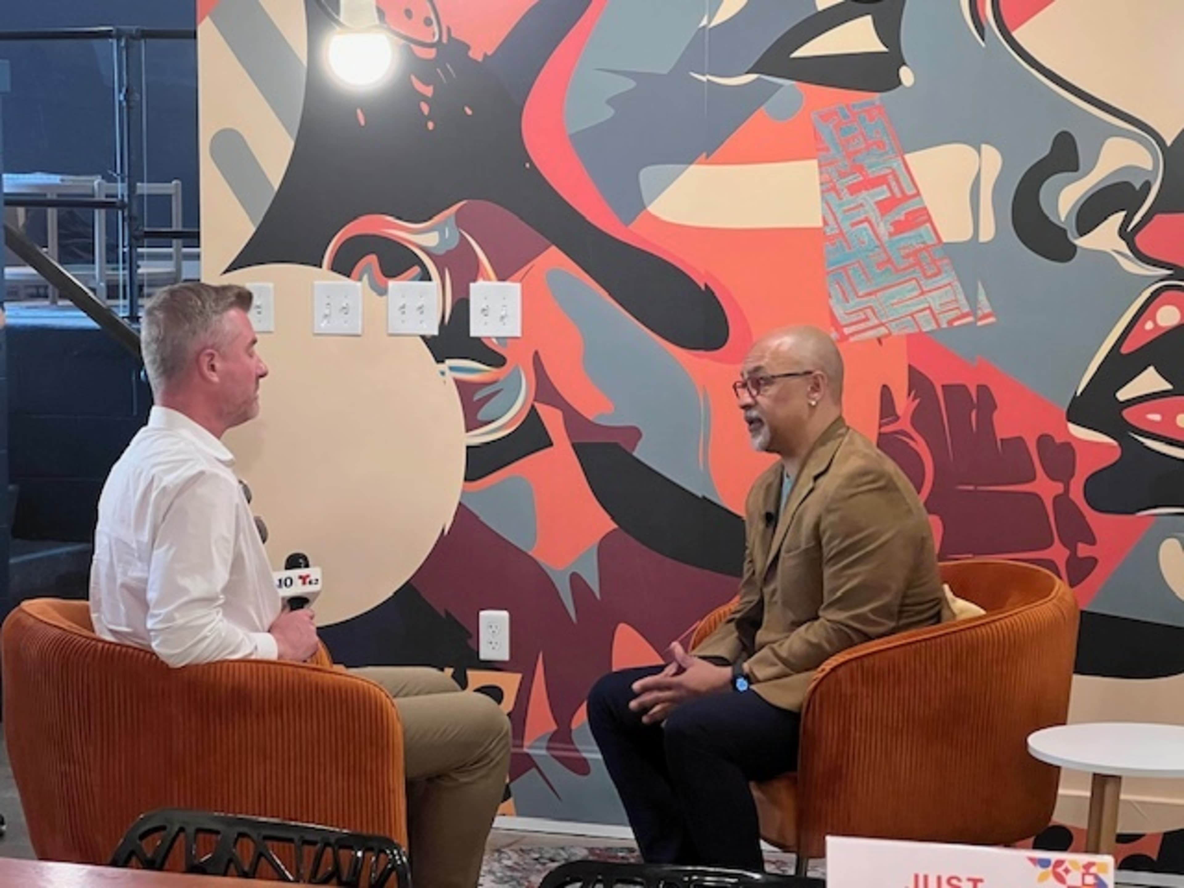 A man in a brown jacket is seated in a chair, engaged in an interview with another man who is holding a microphone, in a room decorated with abstract mural art.