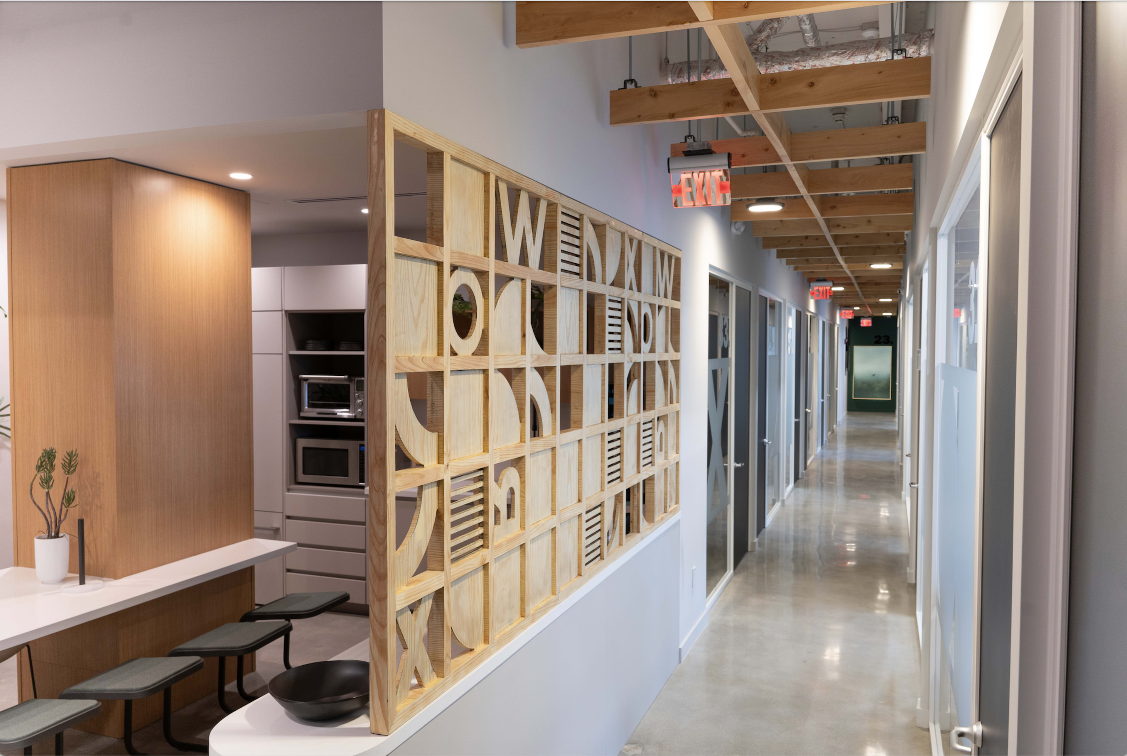 A modern office interior with a wooden partition displaying decorative cutouts, a kitchen area on one side, and a corridor lined with doors on the other.