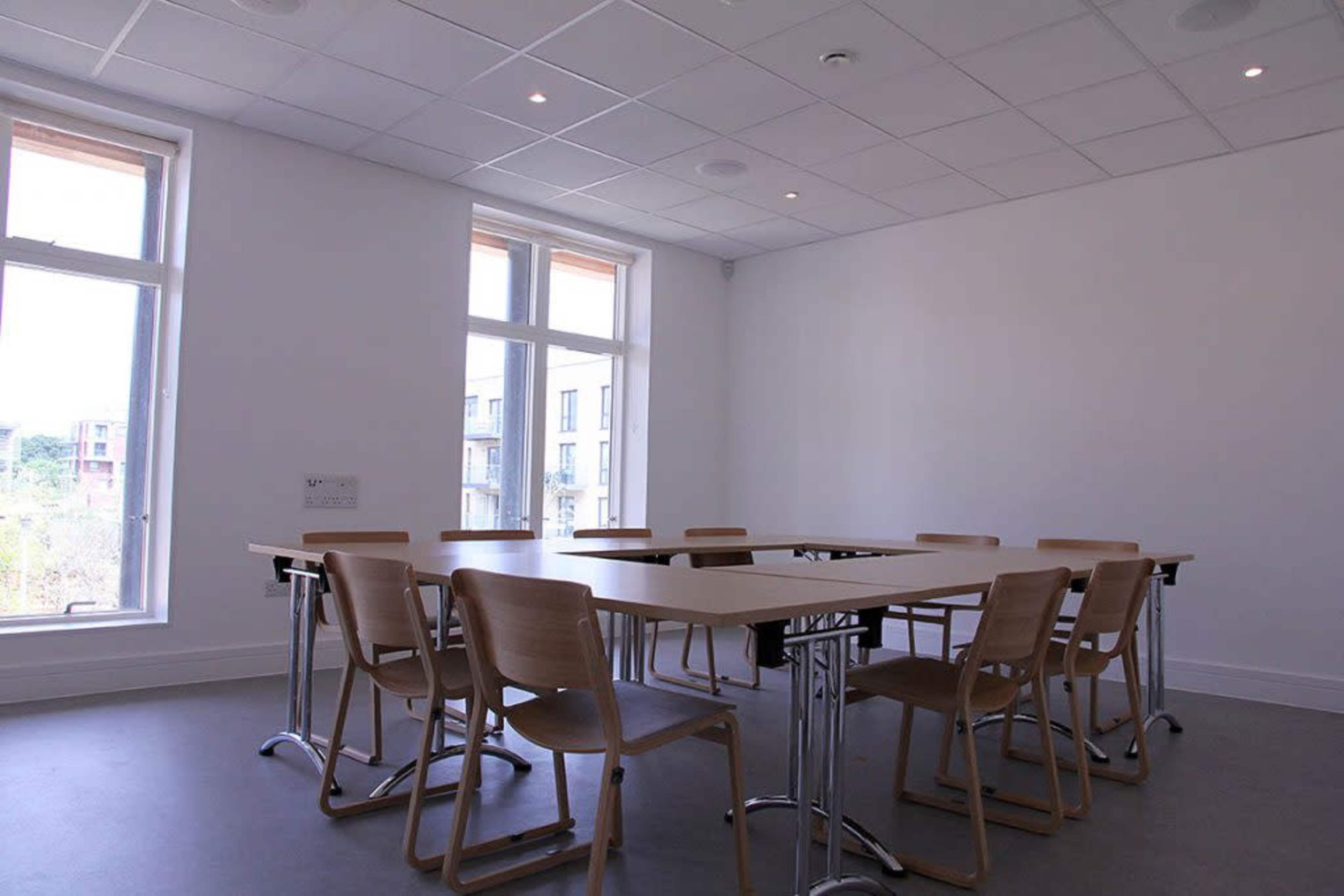 A minimalist conference room features a rectangular table surrounded by eight wooden chairs, with large windows letting in natural light.