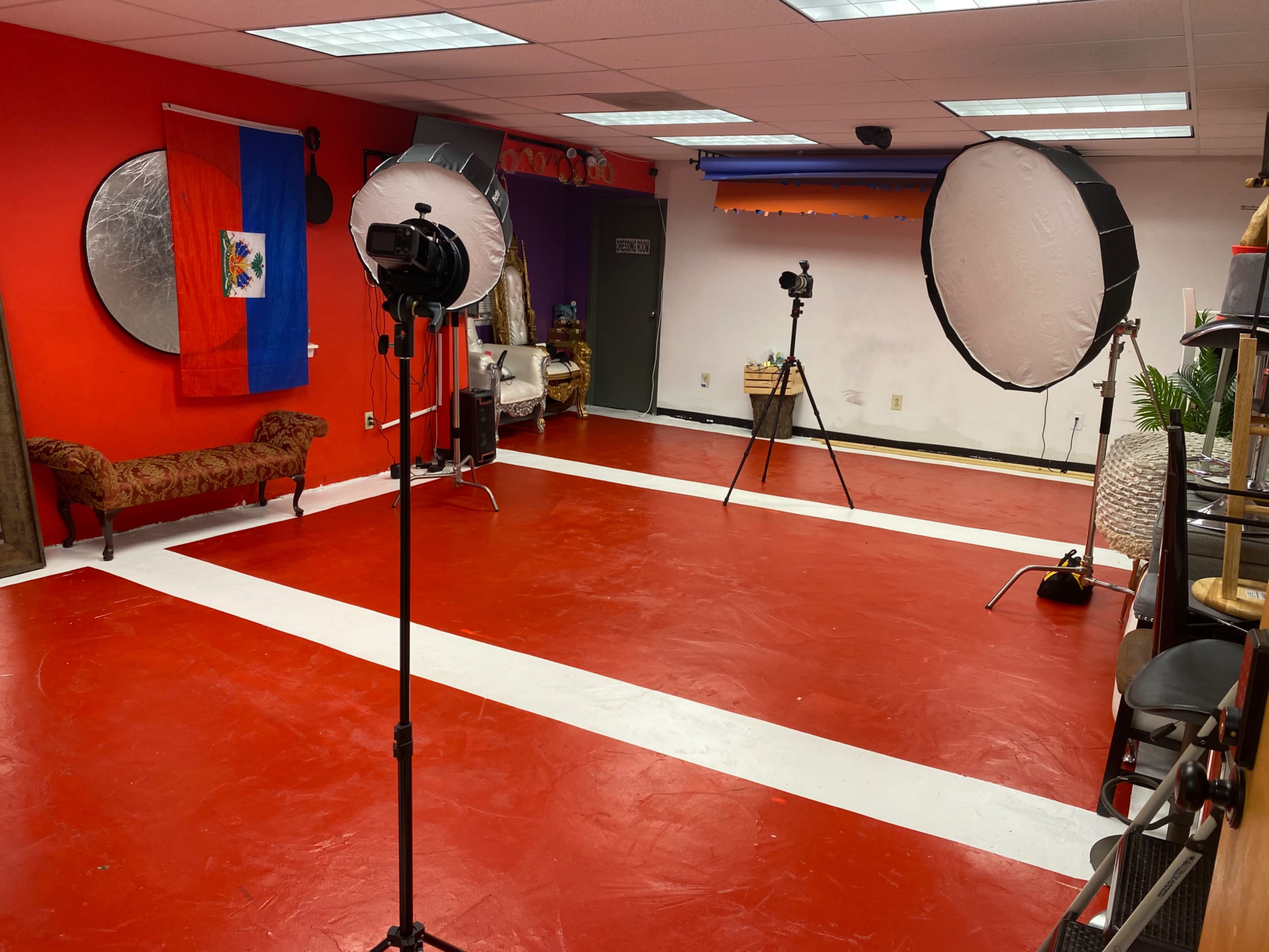 An empty studio space with red flooring, white stripes, two large softbox lights on stands, and a bench, along with a Haitian flag hanging on the wall.