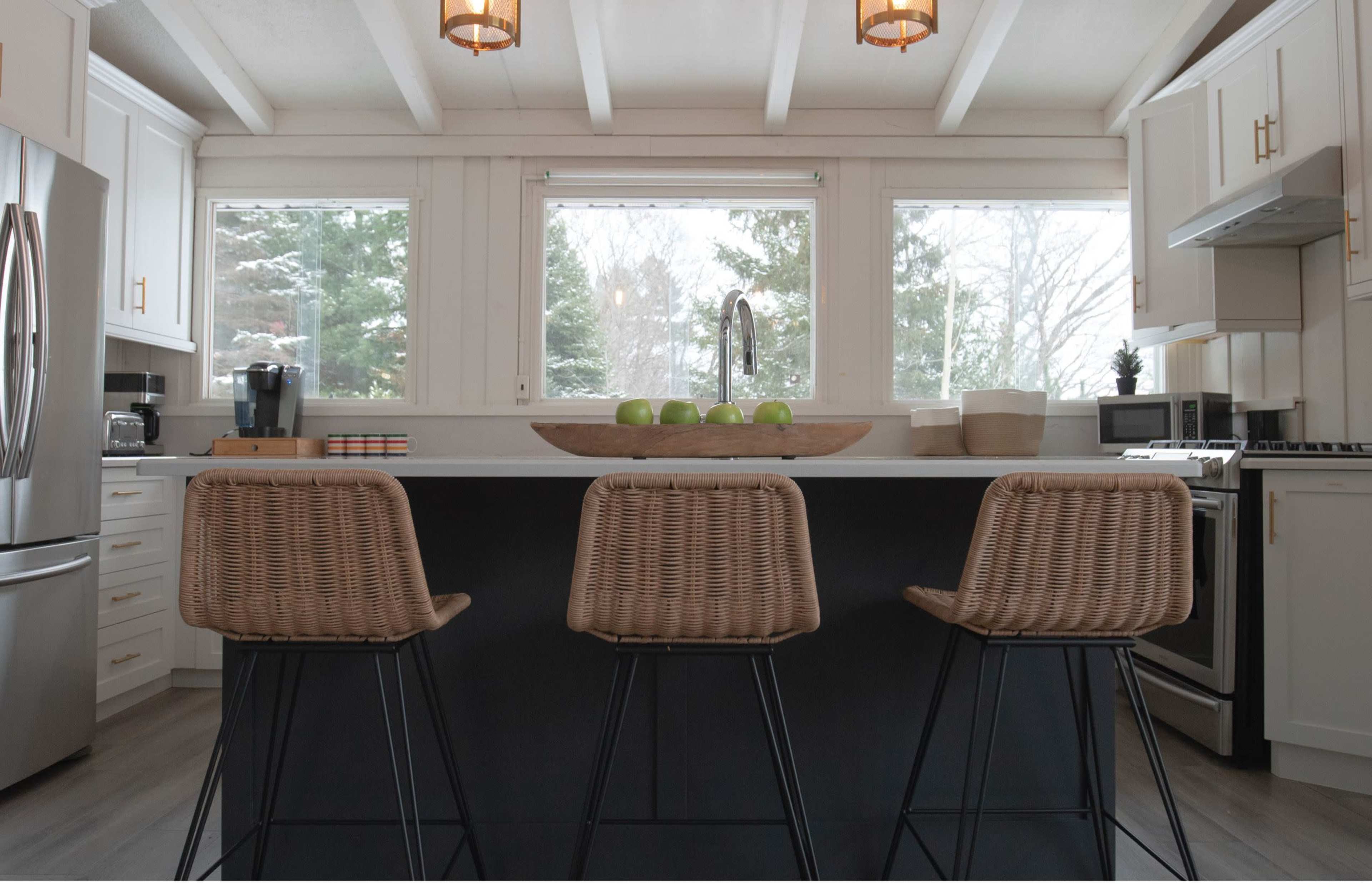 A modern kitchen features an island with three woven bar stools, surrounded by large windows and equipped with stainless steel appliances.