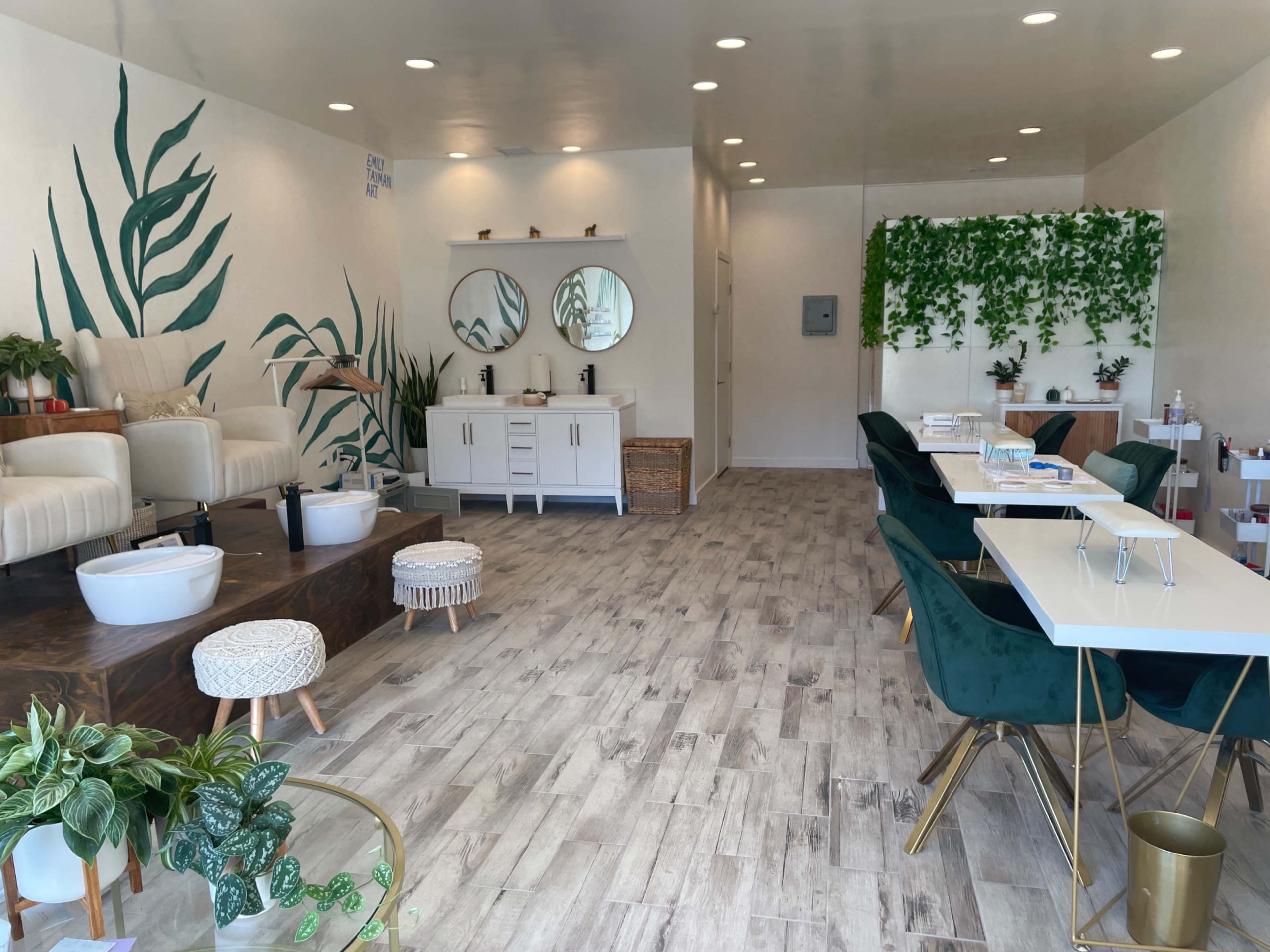 A spa interior featuring modern seating, two mirrored vanities, and greenery along the walls.