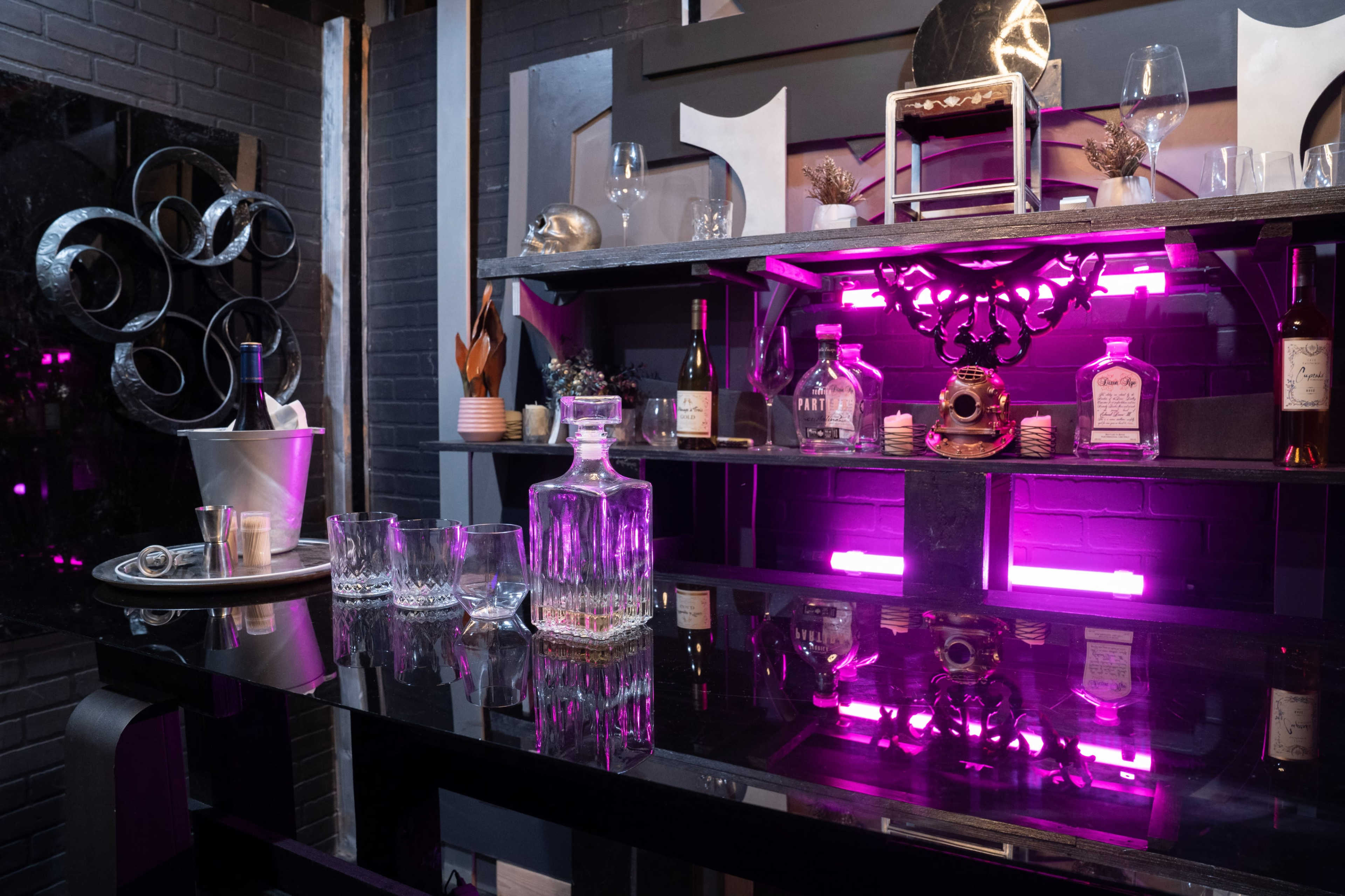 A modern bar setup features a black countertop with a crystal decanter and glasses, alongside various bottles of liquor, illuminated by pink LED lights.