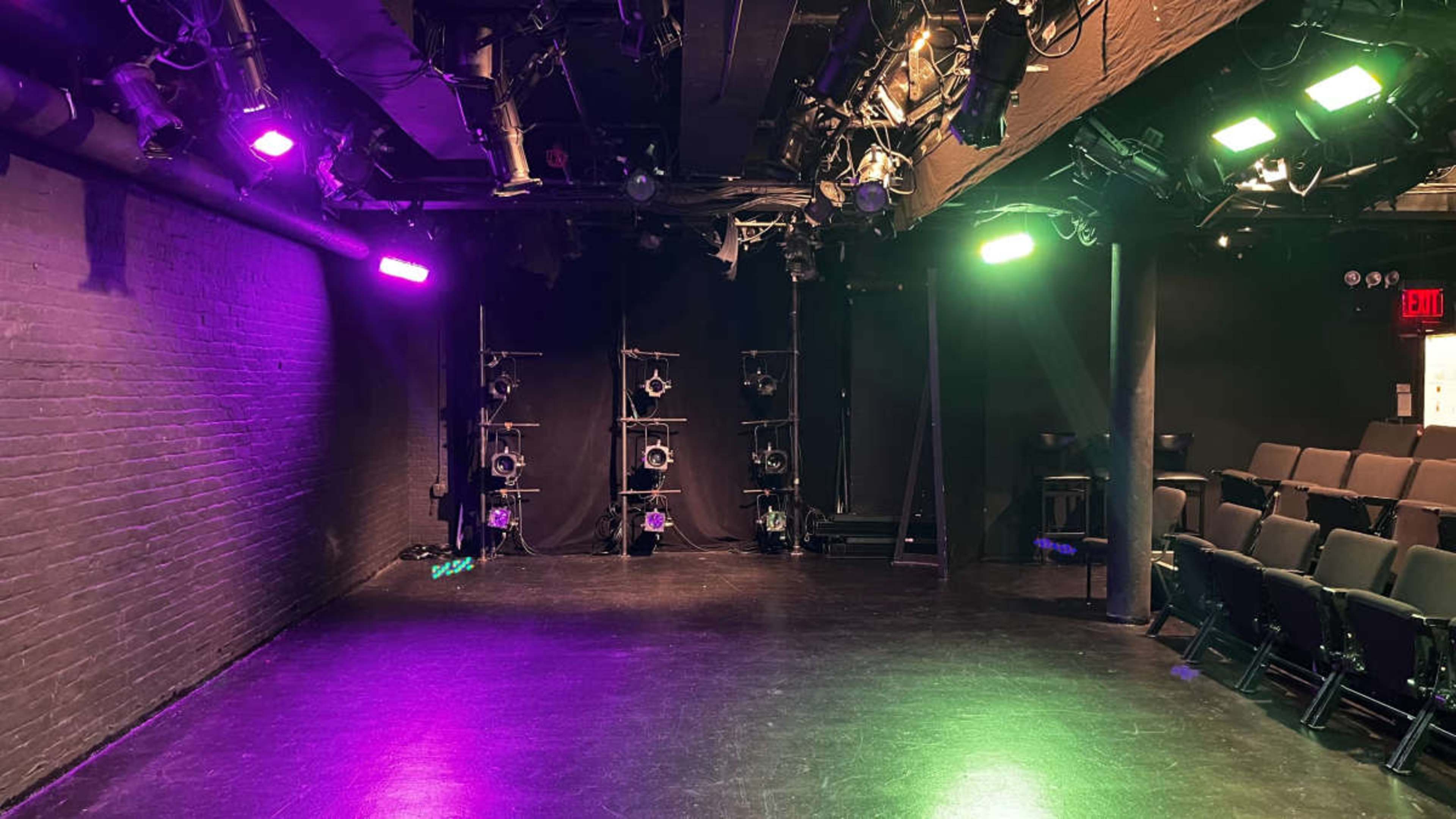 A dimly lit performance space features black walls, seating, and colored stage lights arranged around the area.