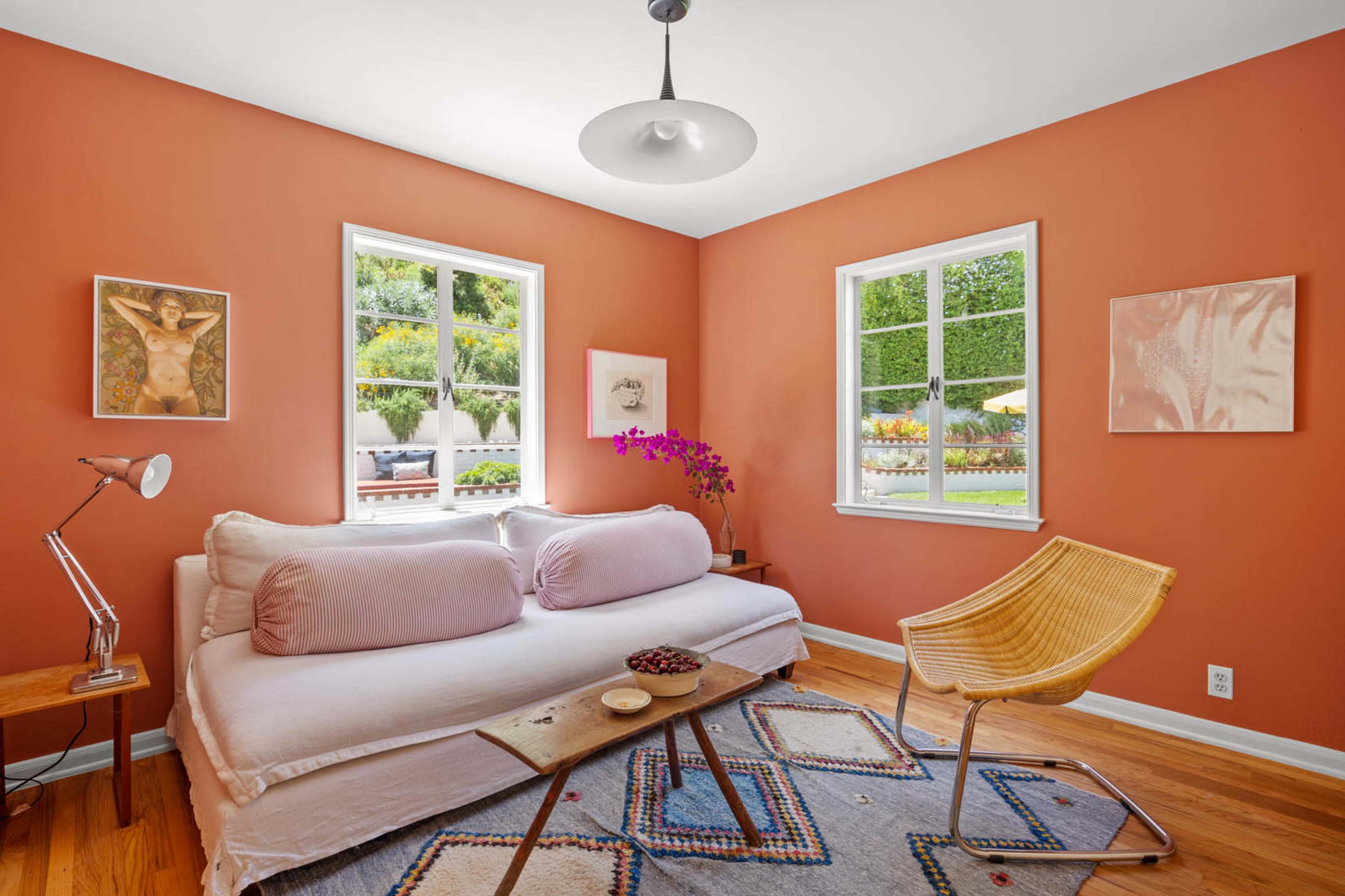 The image shows a cozy bedroom with an orange wall, a daybed adorned with pink pillows, a wooden side table, and large windows letting in natural light.