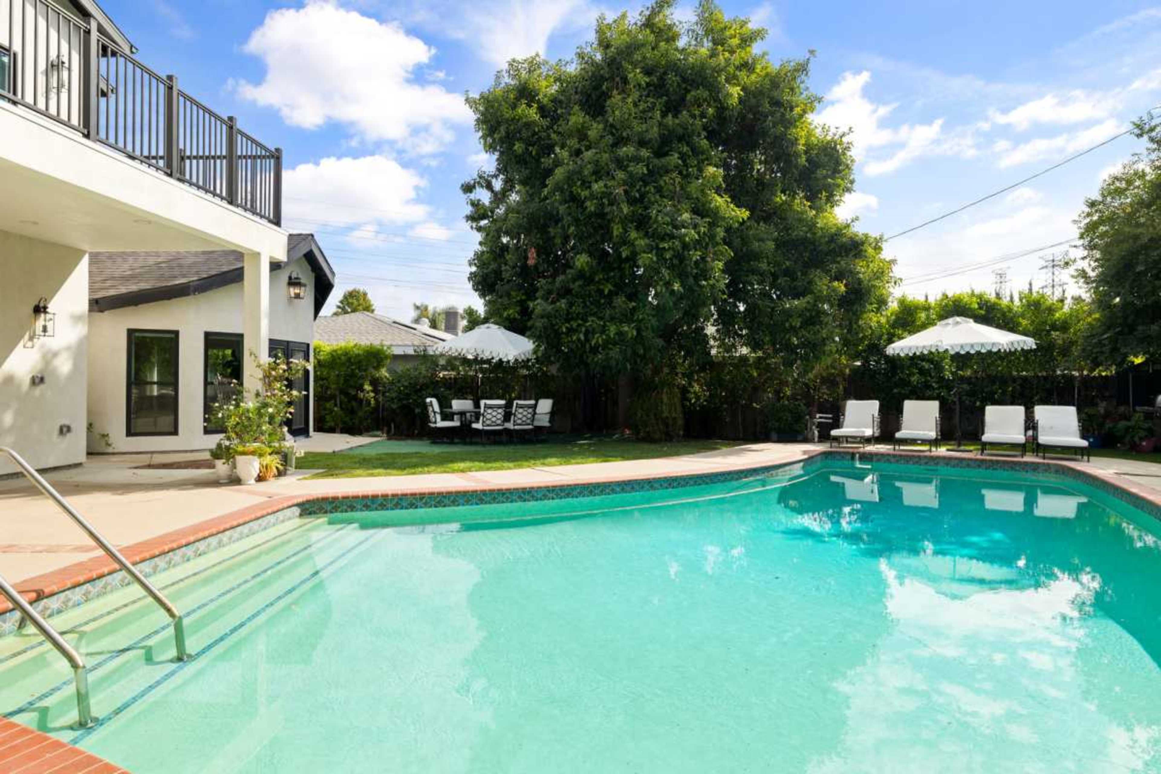A clear blue swimming pool is surrounded by loungers and umbrellas, with a house and greenery in the background.