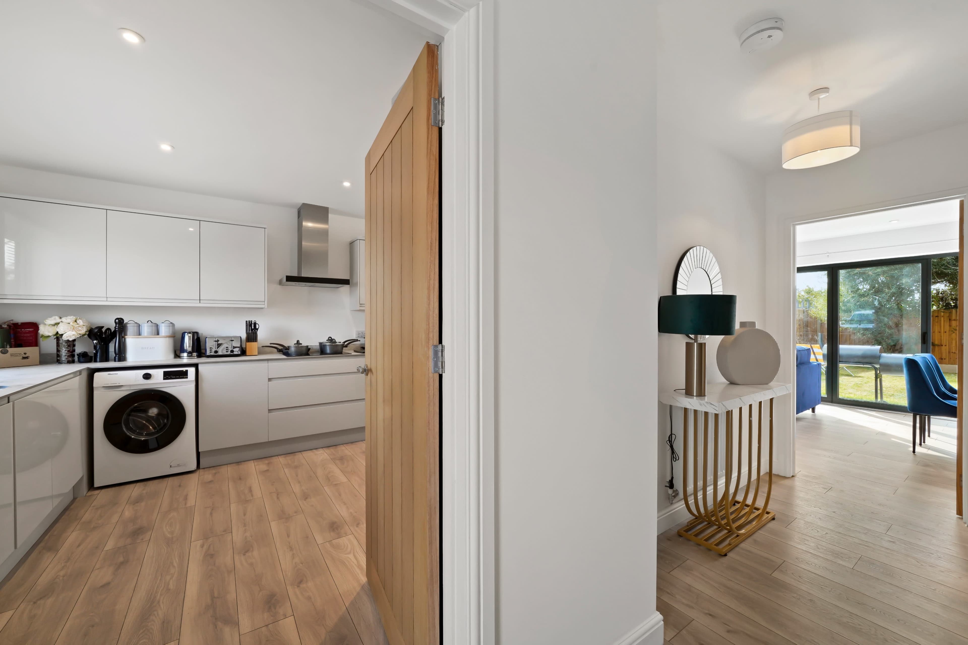 The image shows a modern kitchen with white cabinetry and appliances, adjacent to a hallway that leads to a living area with sliding glass doors.