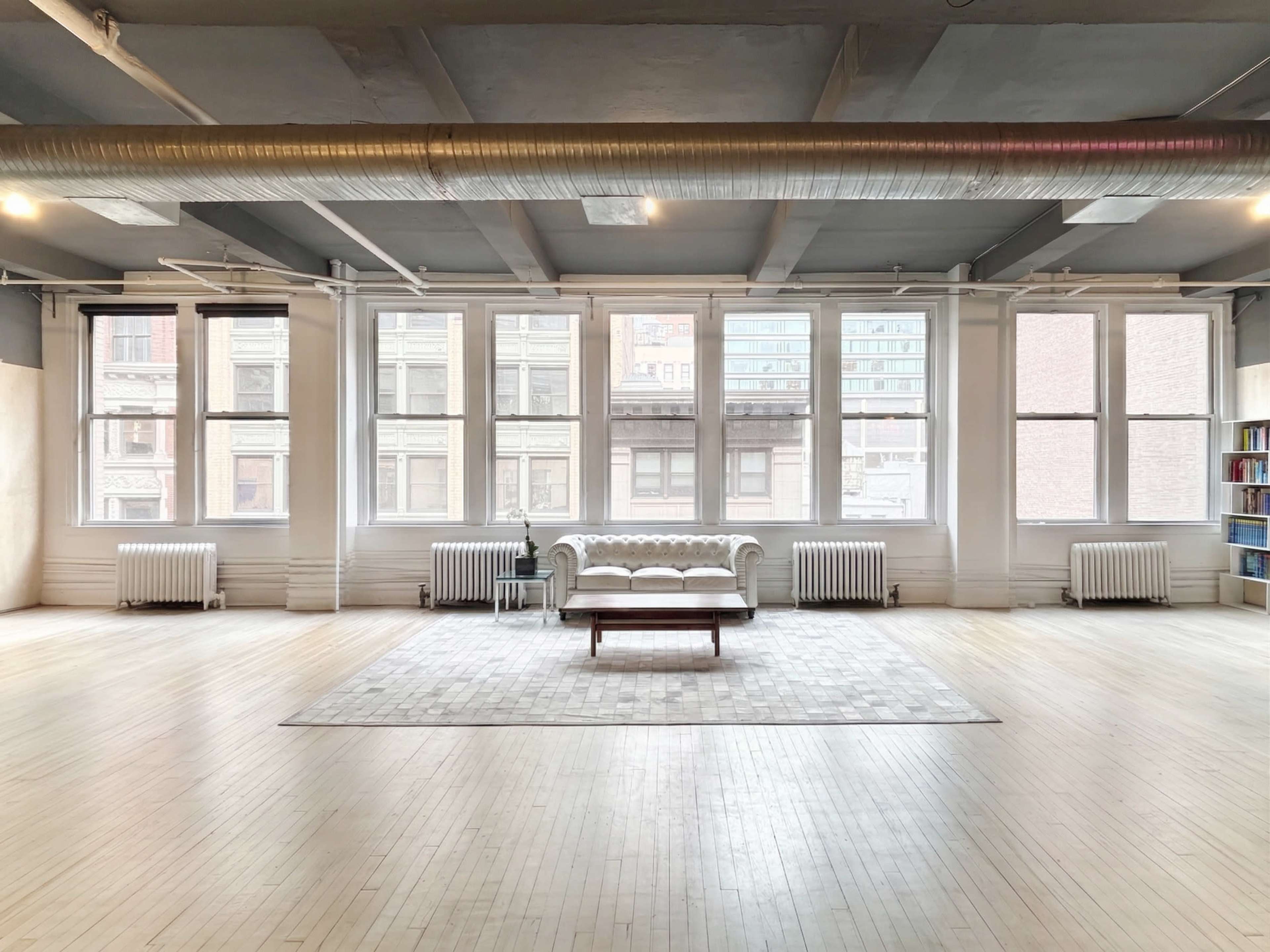 The image shows a spacious room with large windows, a white sofa, a wooden coffee table, and a light-colored rug on a wooden floor.