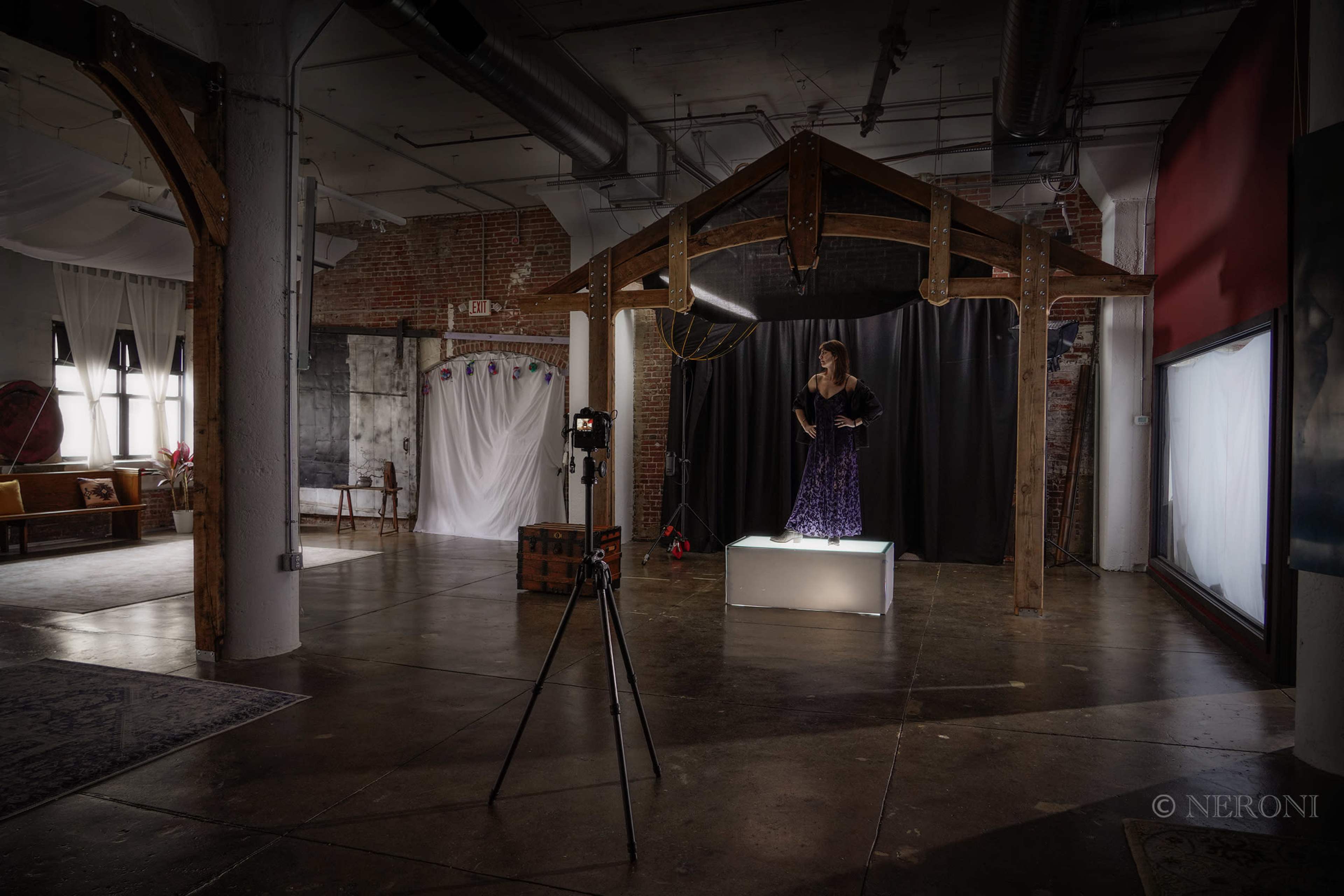 A model poses on a illuminated platform in front of a backdrop while a camera is set up on a tripod in a spacious, industrial setting.