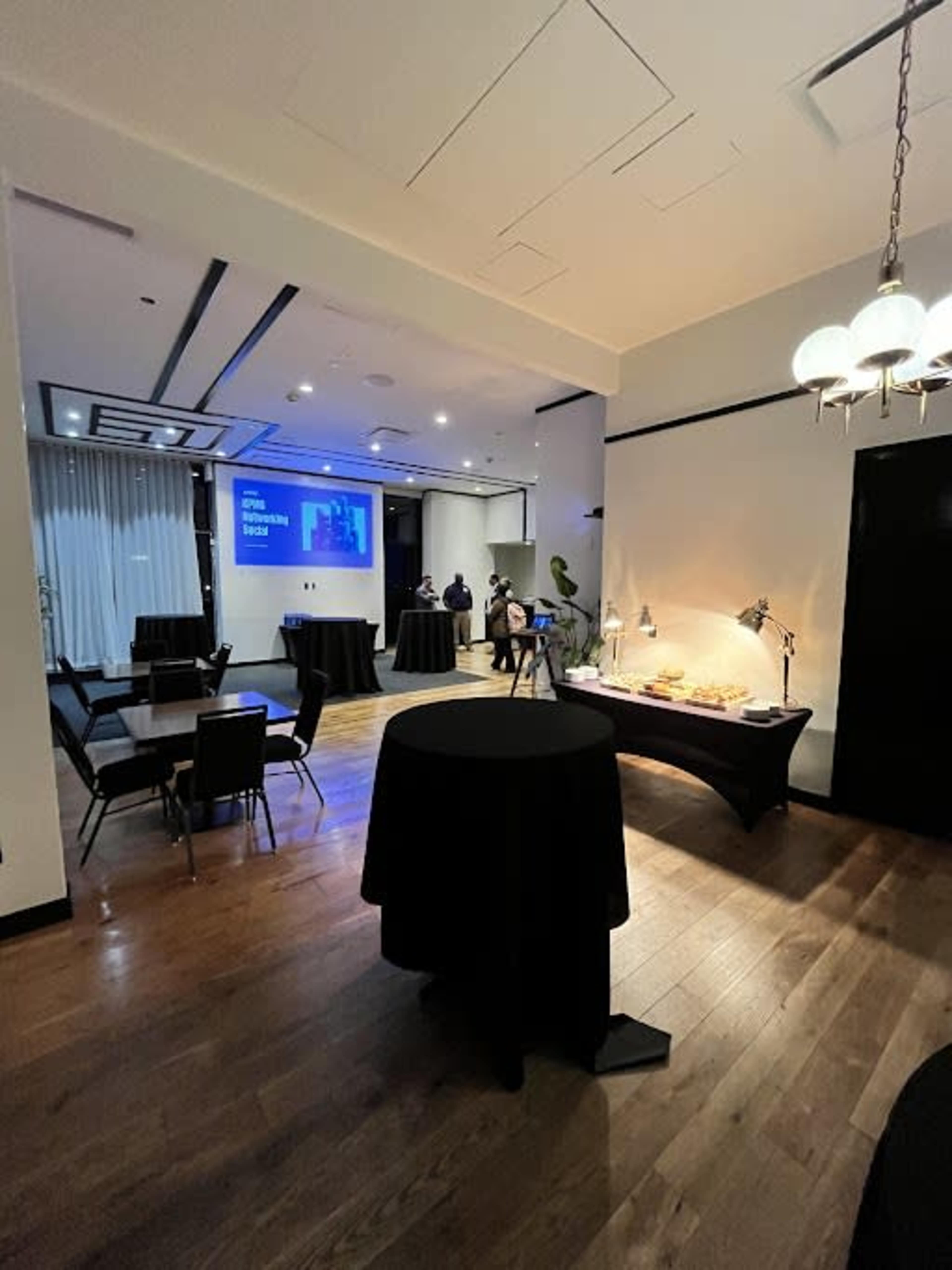 The image shows a spacious event room with black tables, a screen displaying a presentation, and a buffet setup against the wall.