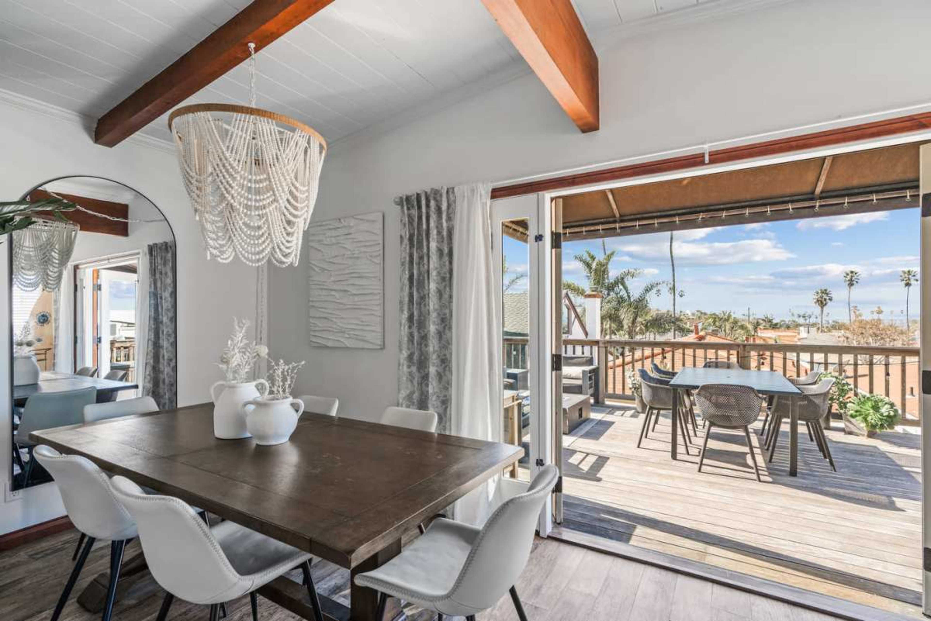 A dining area featuring a wooden table surrounded by white chairs, with large sliding glass doors opening to a deck with a view of palm trees.