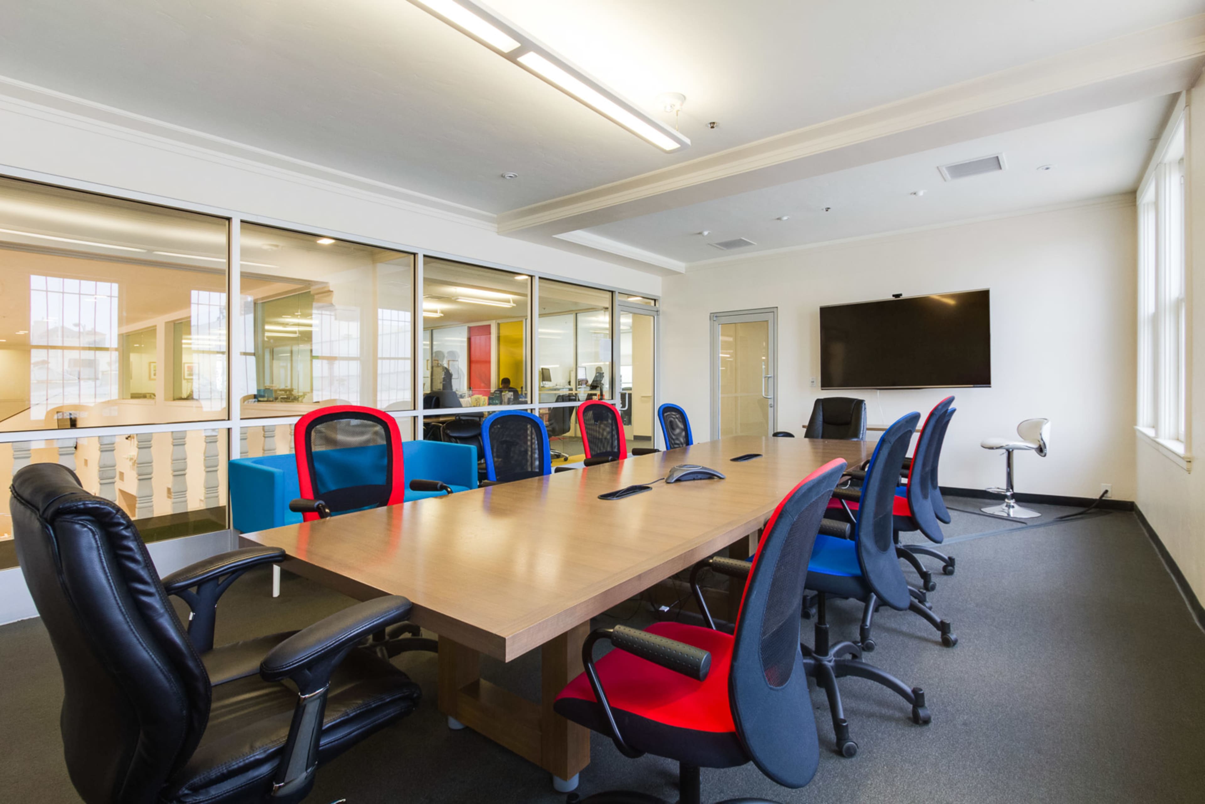A conference room features a large wooden table surrounded by several chairs in different colors, with a wall-mounted television and glass panels offering a view of an adjacent workspace.