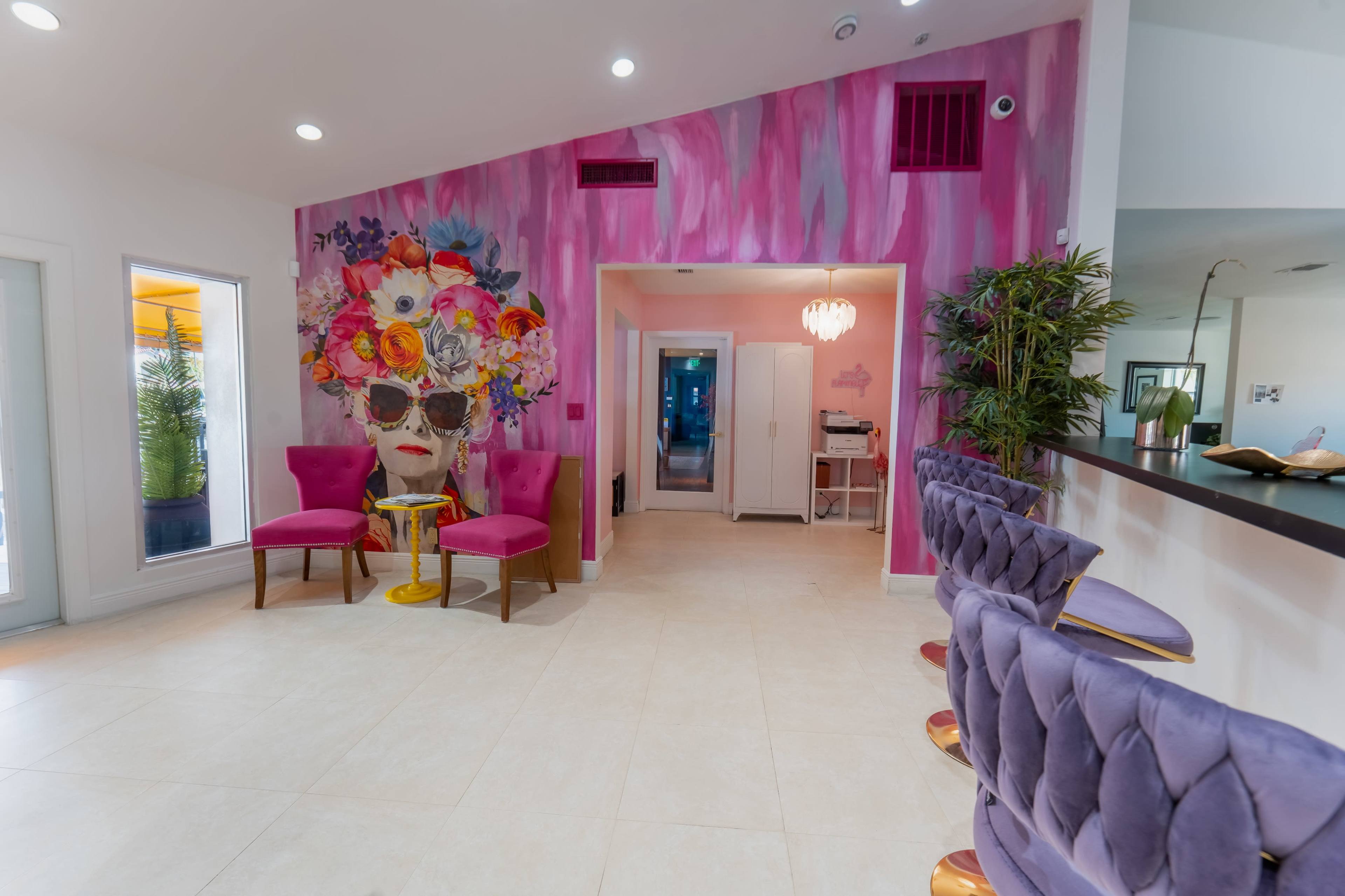 The image shows a modern interior space featuring a vibrant pink wall with a large mural and elegant seating areas, including purple chairs and a bar counter.