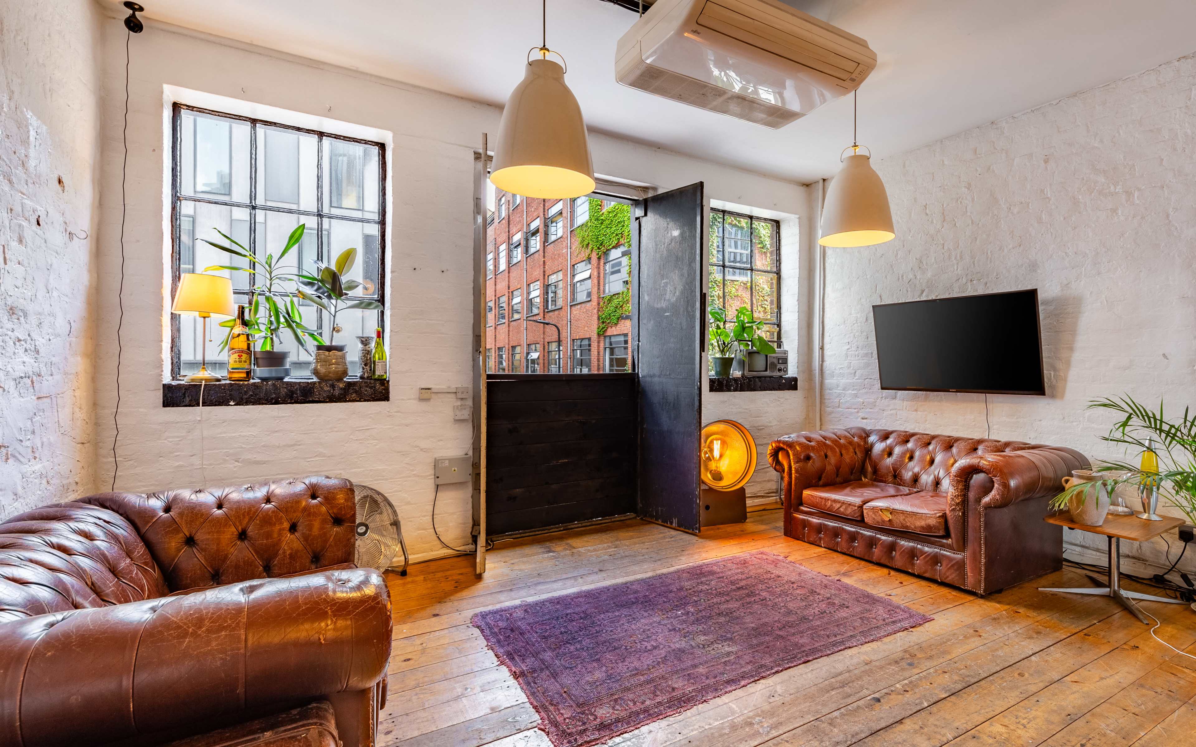 The room features two brown leather sofas, a TV mounted on the wall, and large windows allowing light to stream in, with a door leading outside.