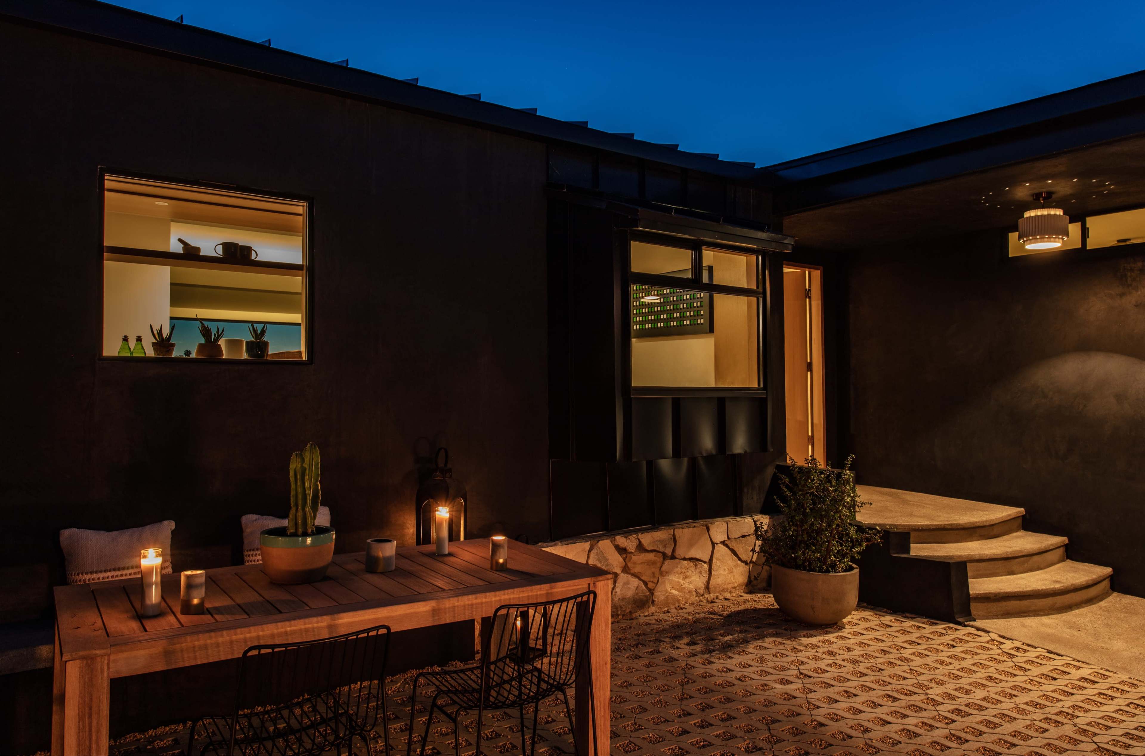 A modern patio features a wooden dining table with candles, surrounded by potted plants, next to a well-lit entrance and stone steps under a twilight sky.