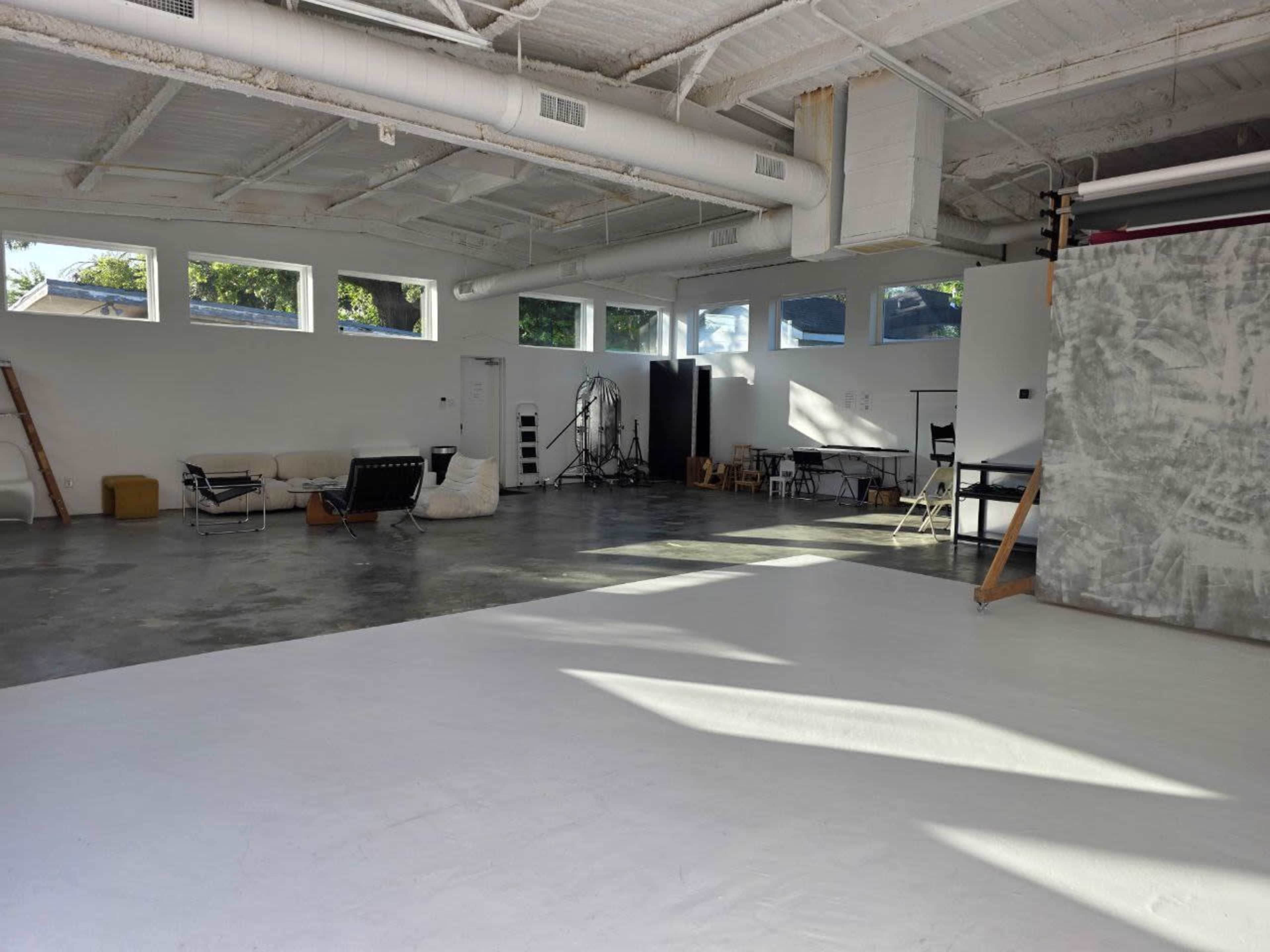The image shows a spacious studio with large windows, a concrete floor, and various furniture pieces arranged around the area.