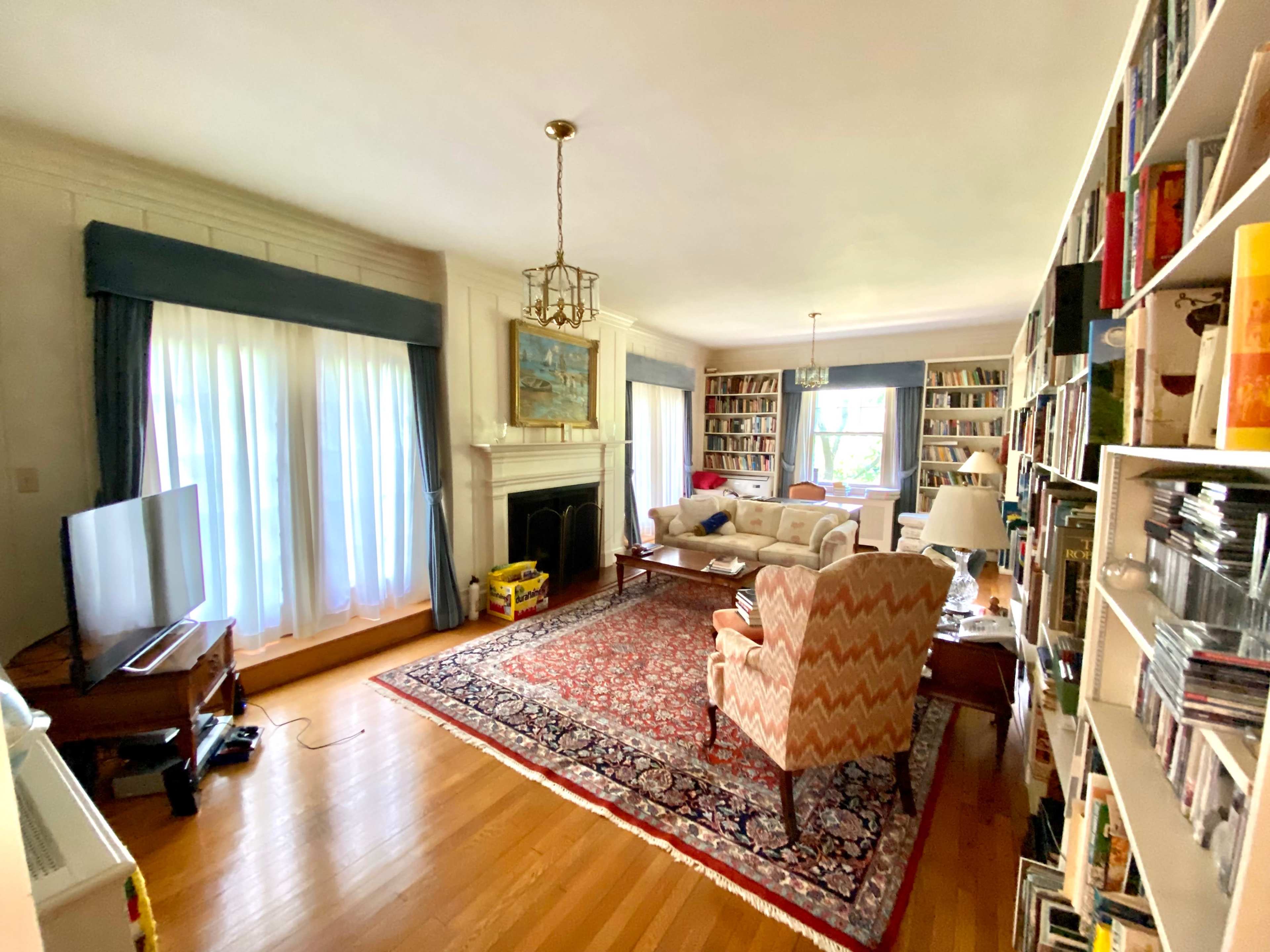 A living room with large windows, a fireplace, bookshelves filled with books, and a seating area featuring a sofa and chairs.