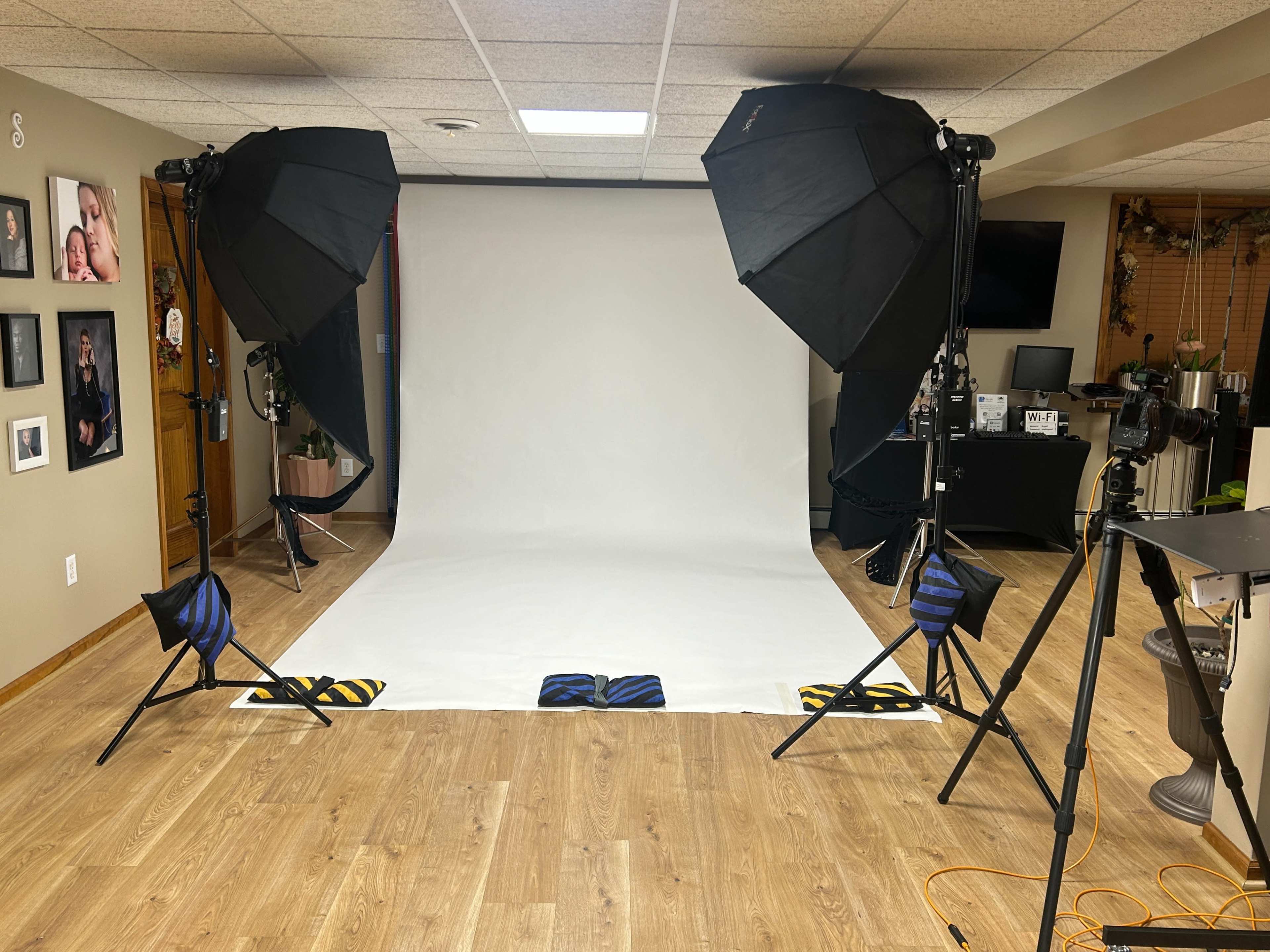 The image shows a photography studio setup with two large softbox lights, a white backdrop, and a camera on a tripod in a room with wood flooring.