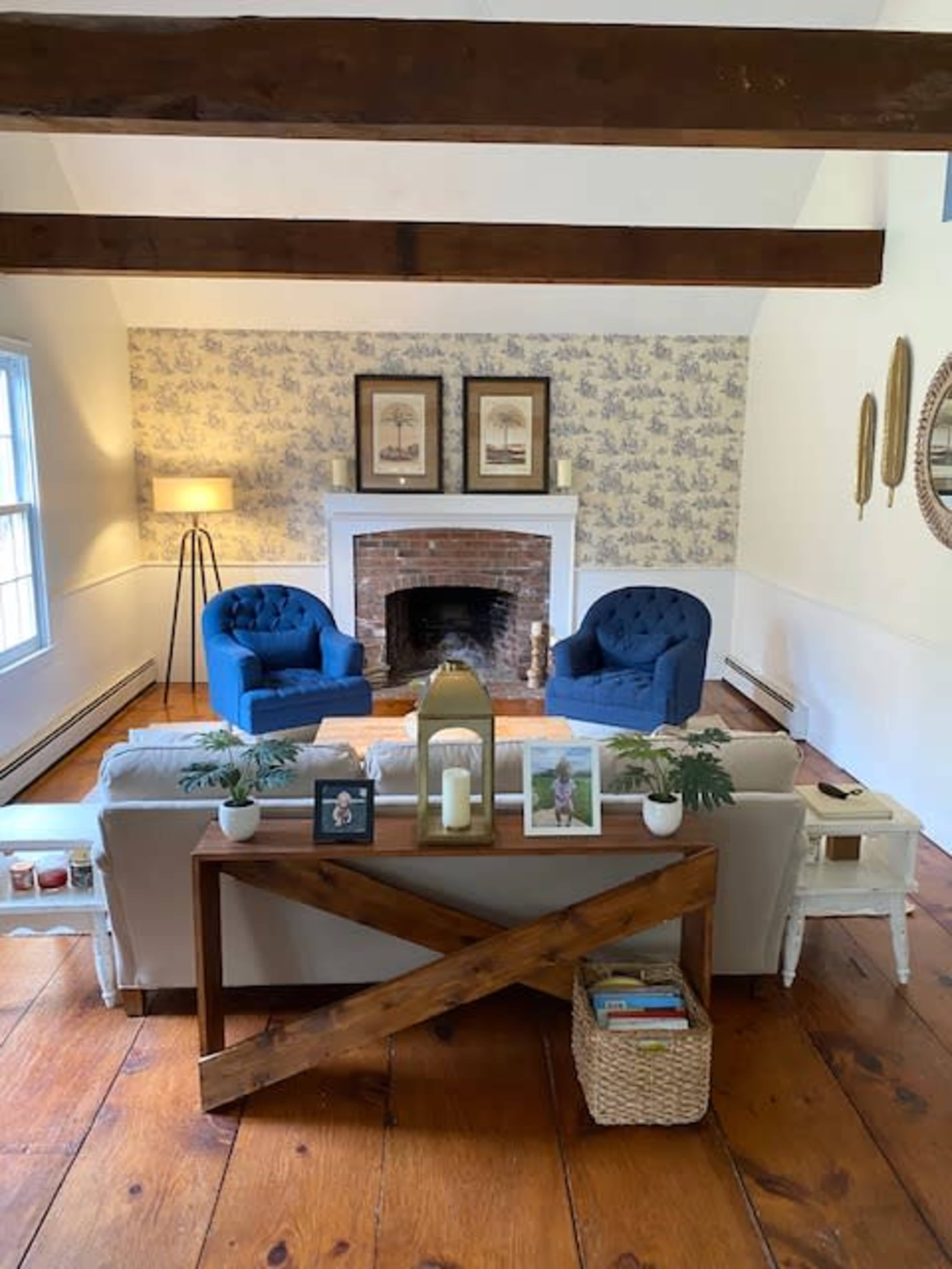 The image shows a cozy living room with two blue upholstered chairs facing a brick fireplace, a white couch, and a wooden console table with decorative items.