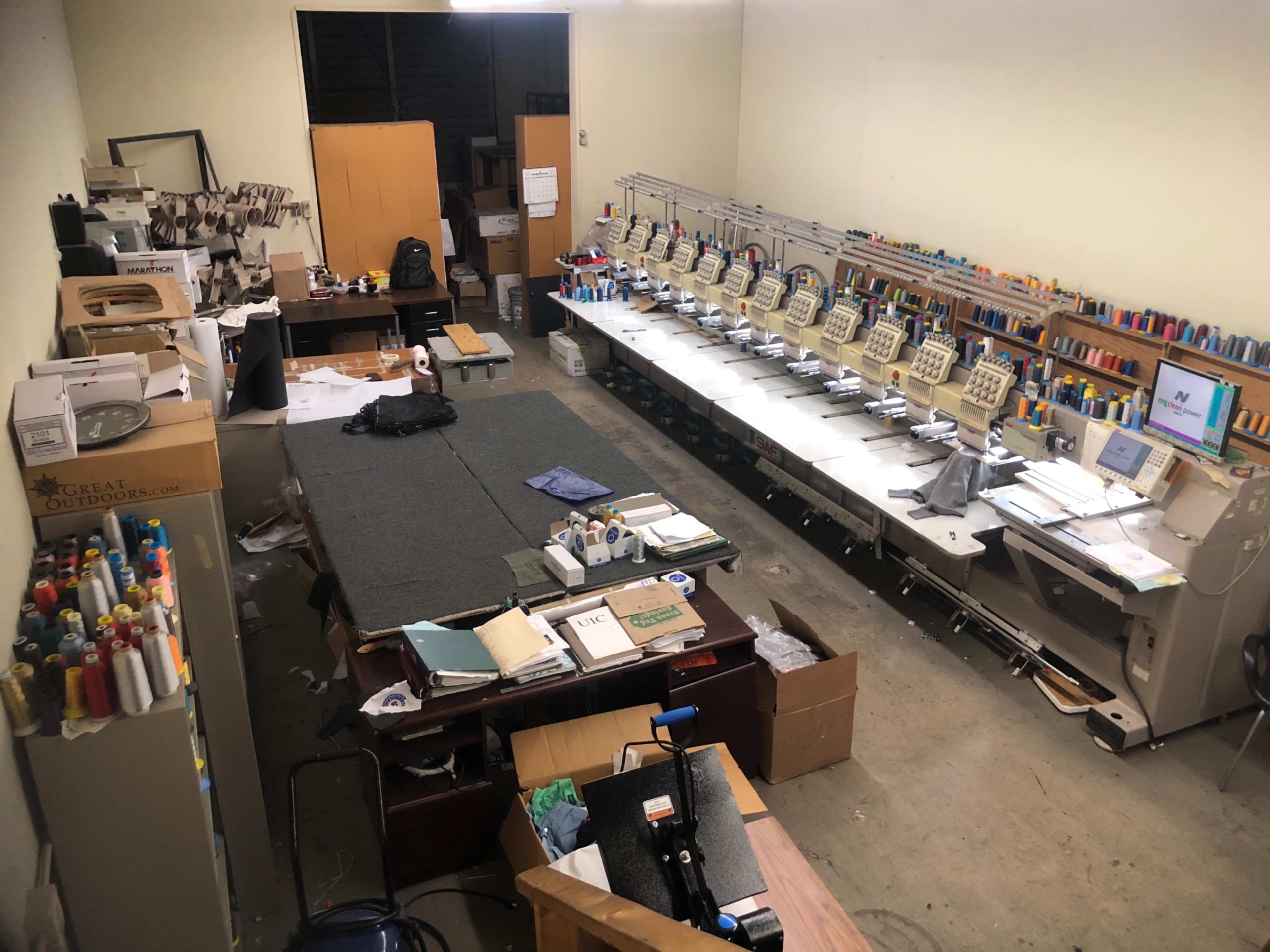 A cluttered sewing and embroidery workspace with multiple embroidery machines lined up along one side and scattered materials, papers, and tools throughout the room.