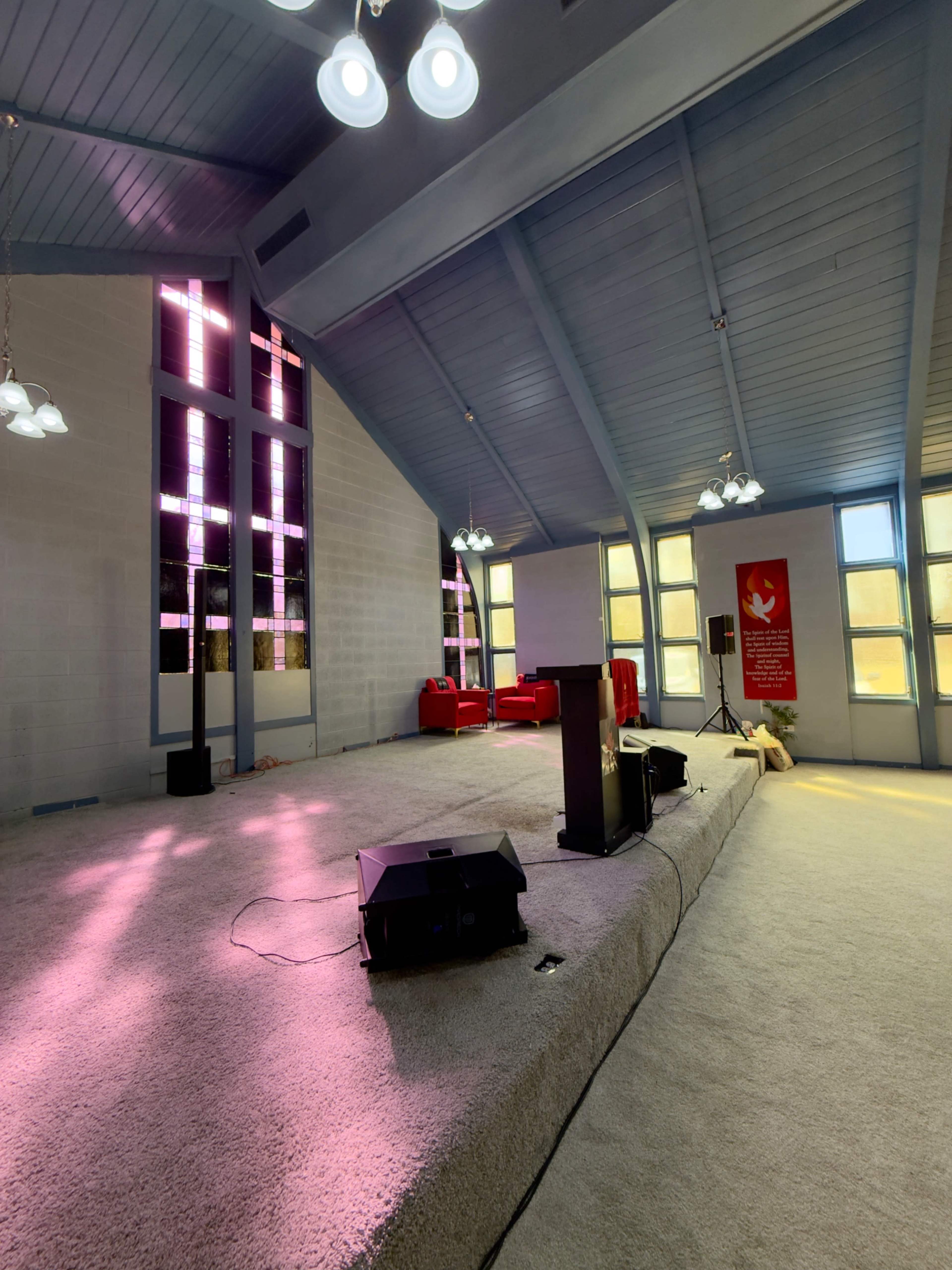 The interior of a church features a raised platform with a microphone stand and speakers, surrounded by colorful stained glass windows and red seating.