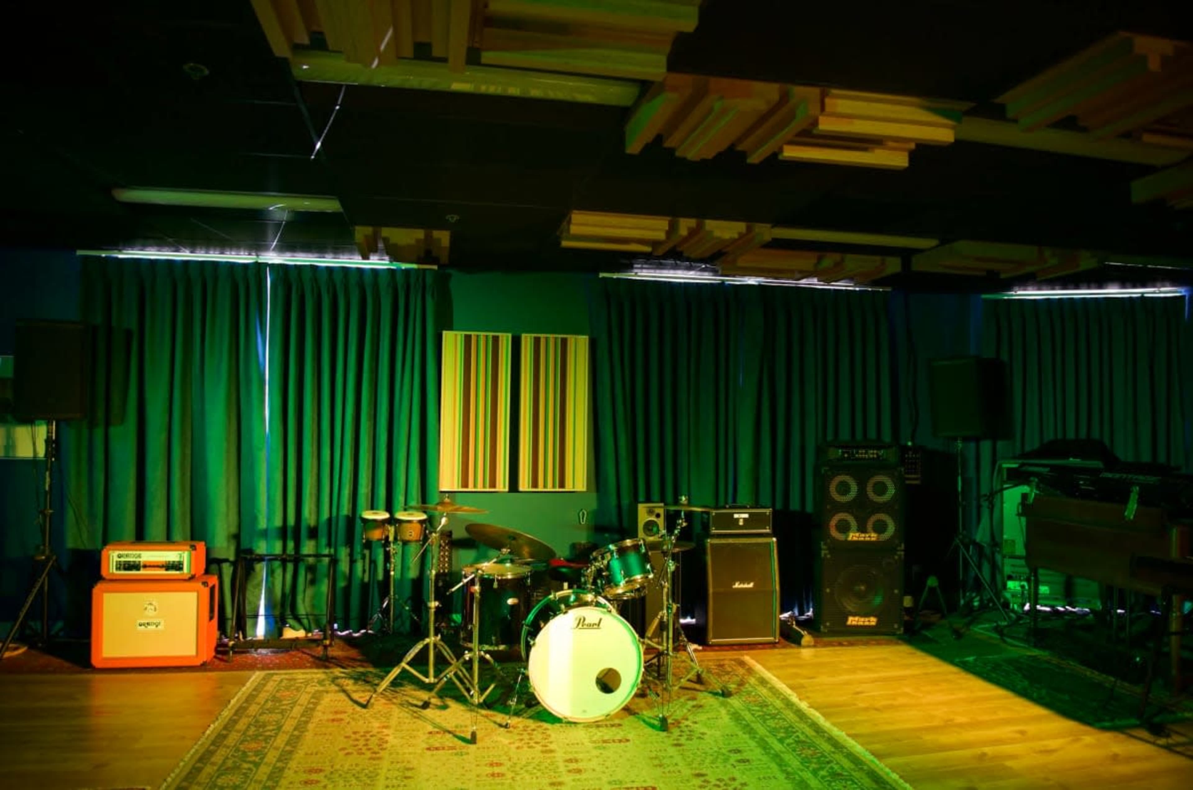 A music studio is set up with a drum kit, amplifiers, and sound equipment, surrounded by green curtains and acoustic panels.