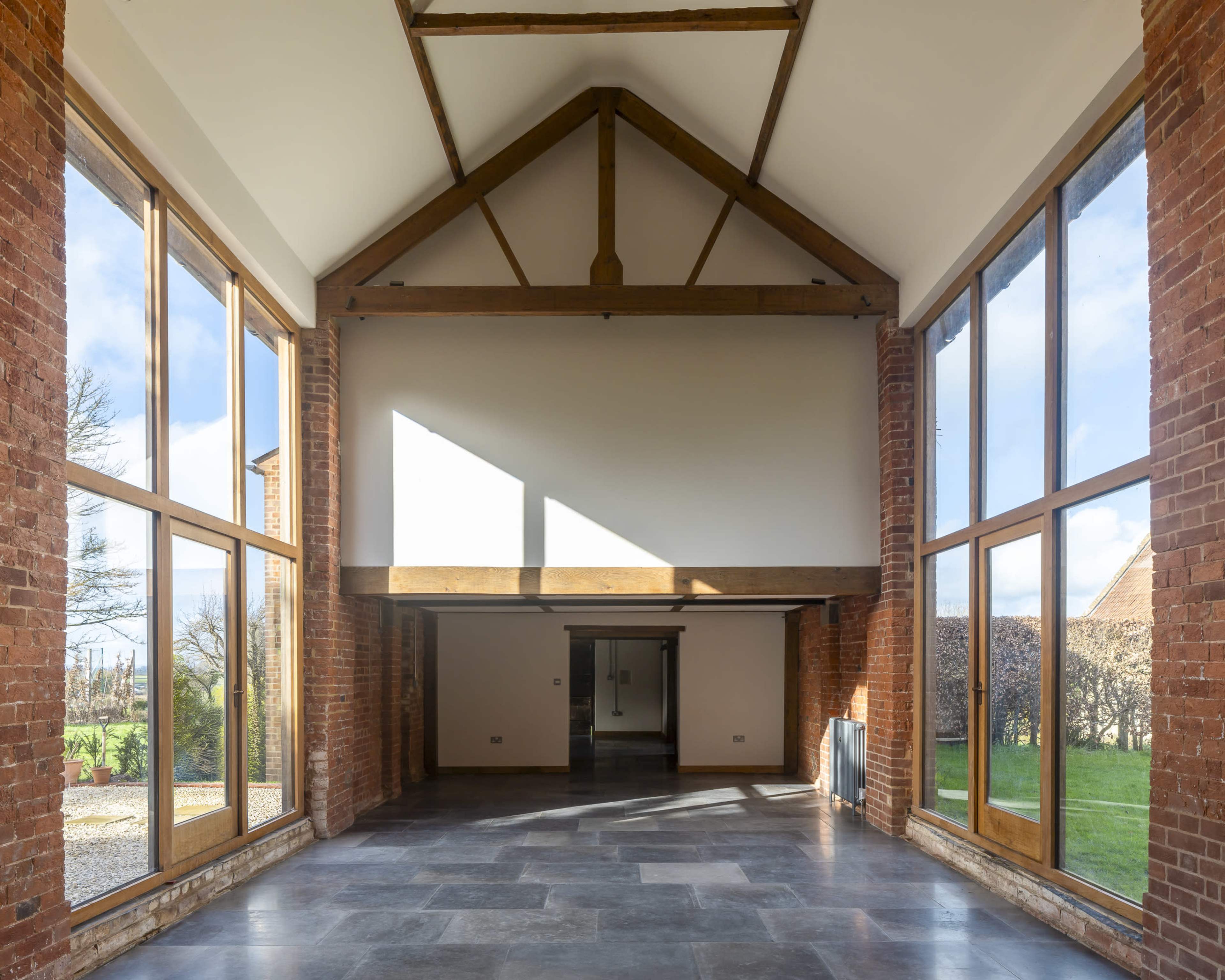The image shows a spacious interior with large windows, exposed wooden beams, and a stone floor, providing views of a green landscape outside.